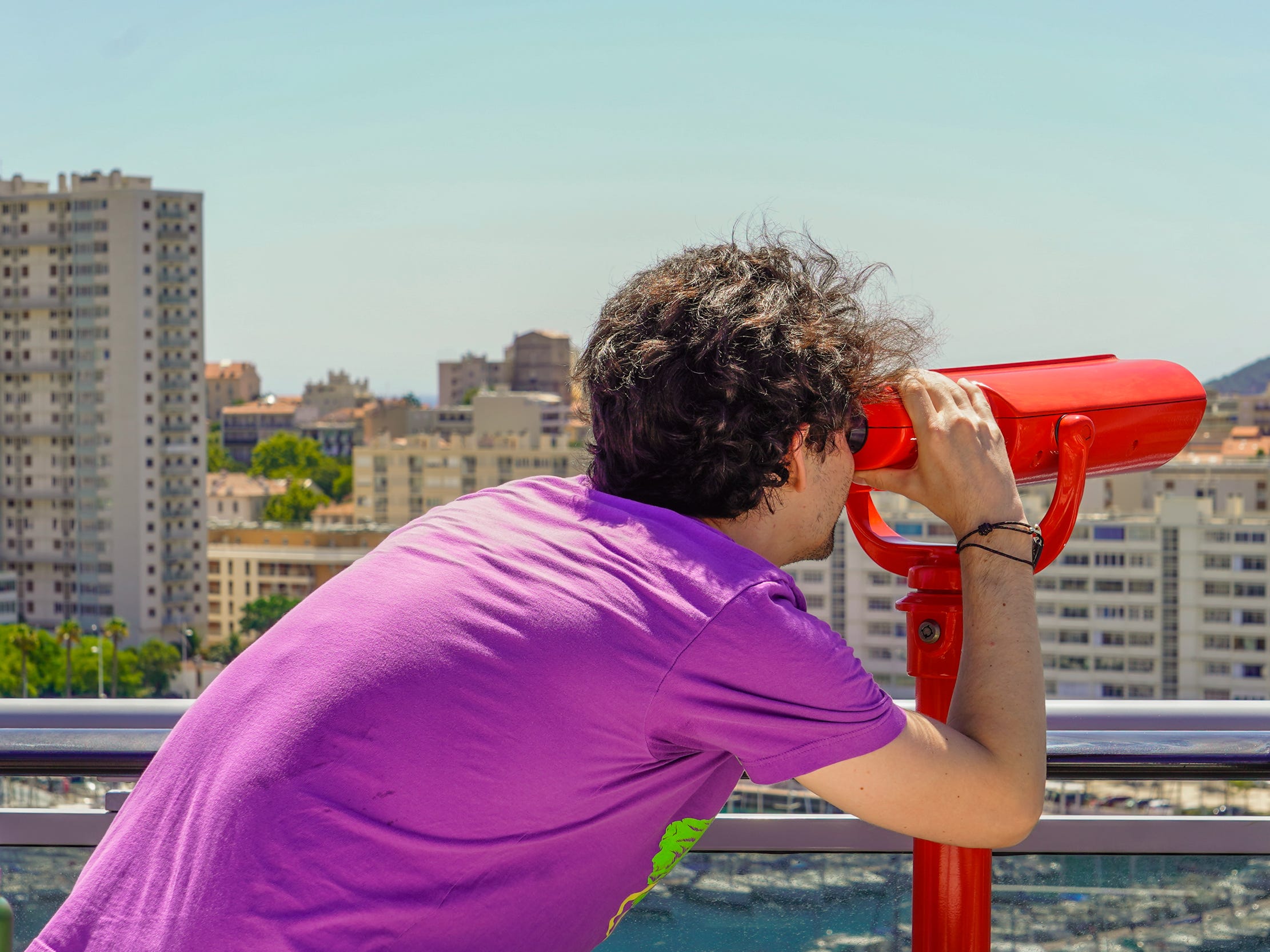 Ein Mann in einem lila Hemd blickt durch ein rotes Teleskop auf Gebäude an einer Küste vor sich