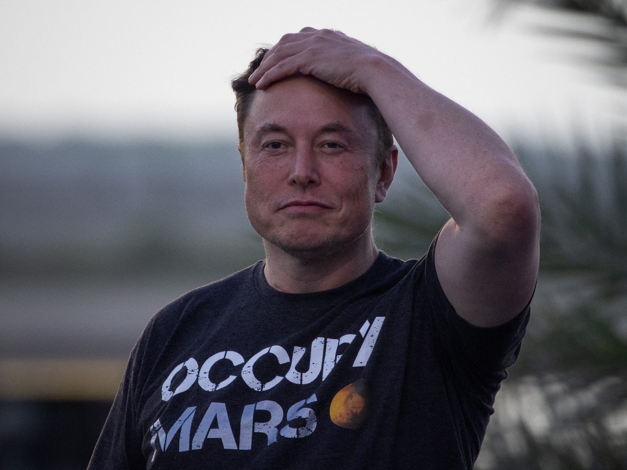 Elon Musk runzelt die Stirn, grinst mit der Hand auf dem Kopf und trägt ein schwarzes Occupy-Mars-Shirt