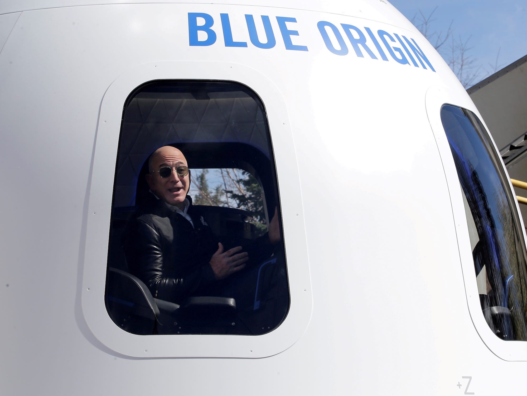 Jeff Bezos mit Glatze und Sonnenbrille gestikuliert mit offenem Mund im Fenster einer Raumkapsel von Blue Origin