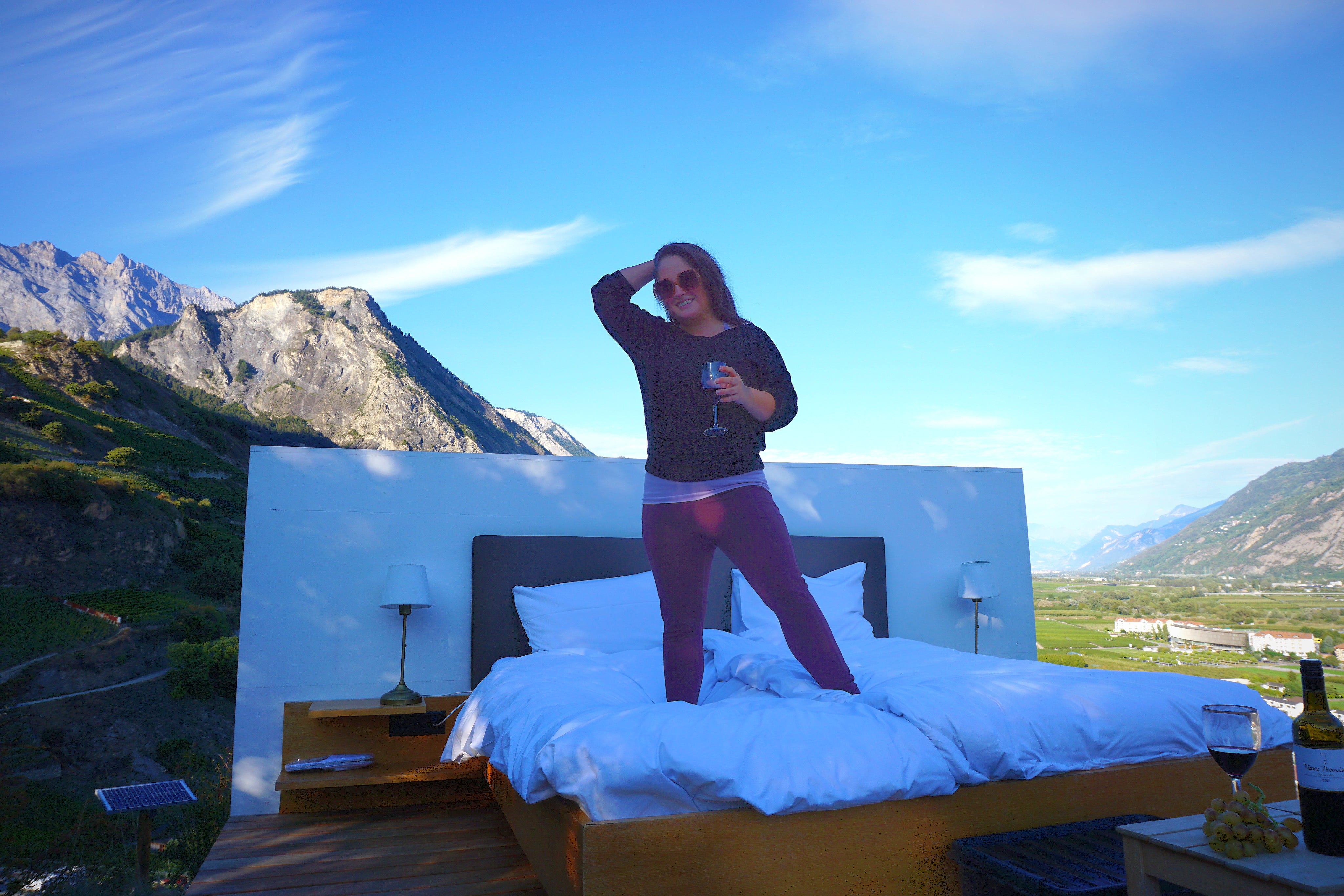 Katka Reversosova hält ein Weinglas in der Hand und steht auf einem Bett vor den Bergen in der Schweiz
