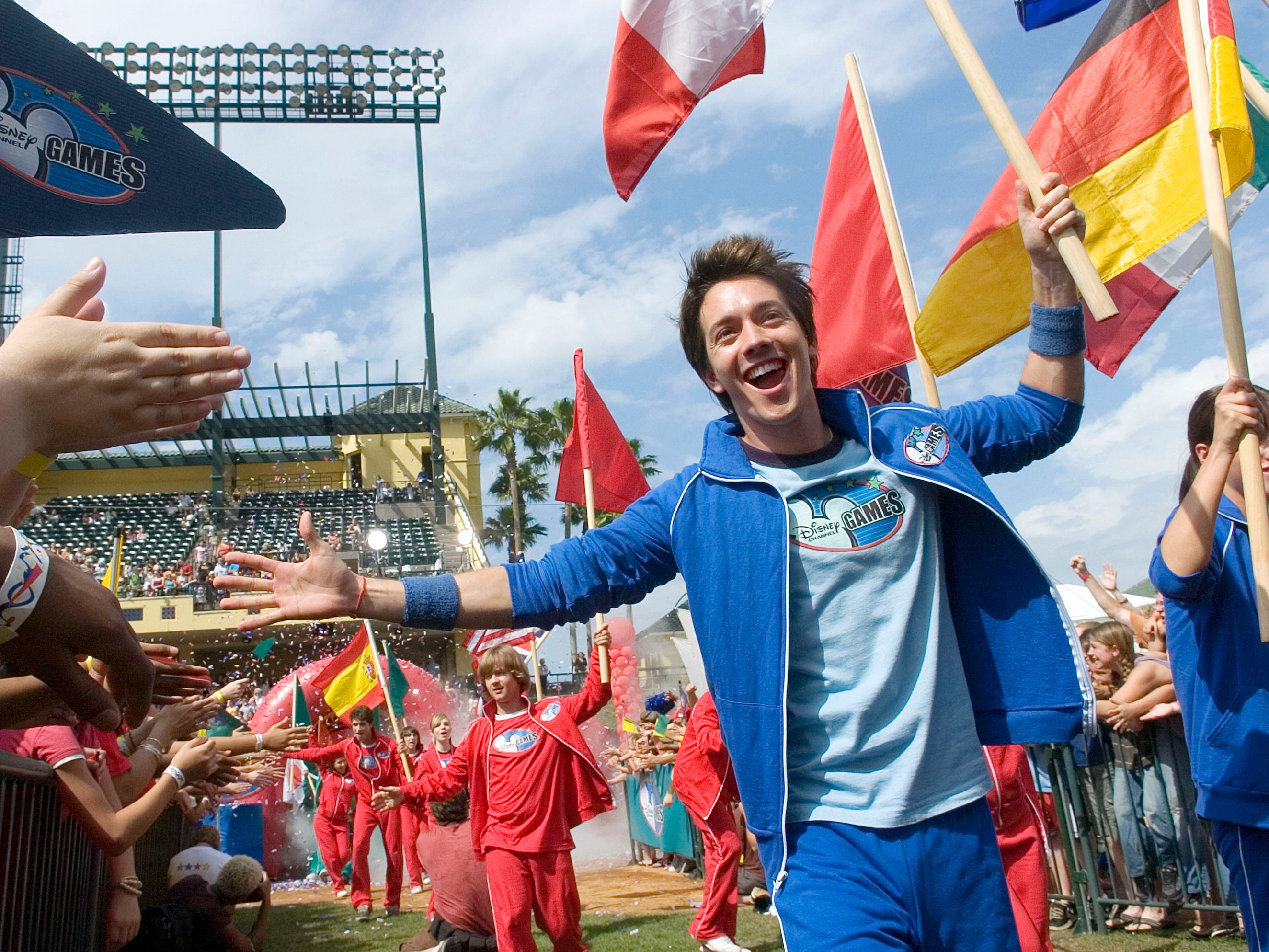 Roger Gonzalez-Garca trägt bei den Disney-Channel-Spielen im Jahr 2007 einen blauen Trainingsanzug mit einer Hand, die er den High-Five-Fans entgegenstreckt, und einer Flagge in der anderen Hand