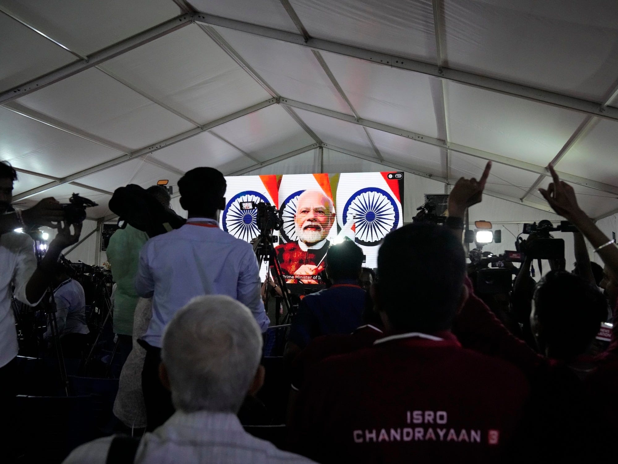 Ein Raum voller Männer beobachtet Narendra Modi auf einem Bildschirm unter einer weißen Decke