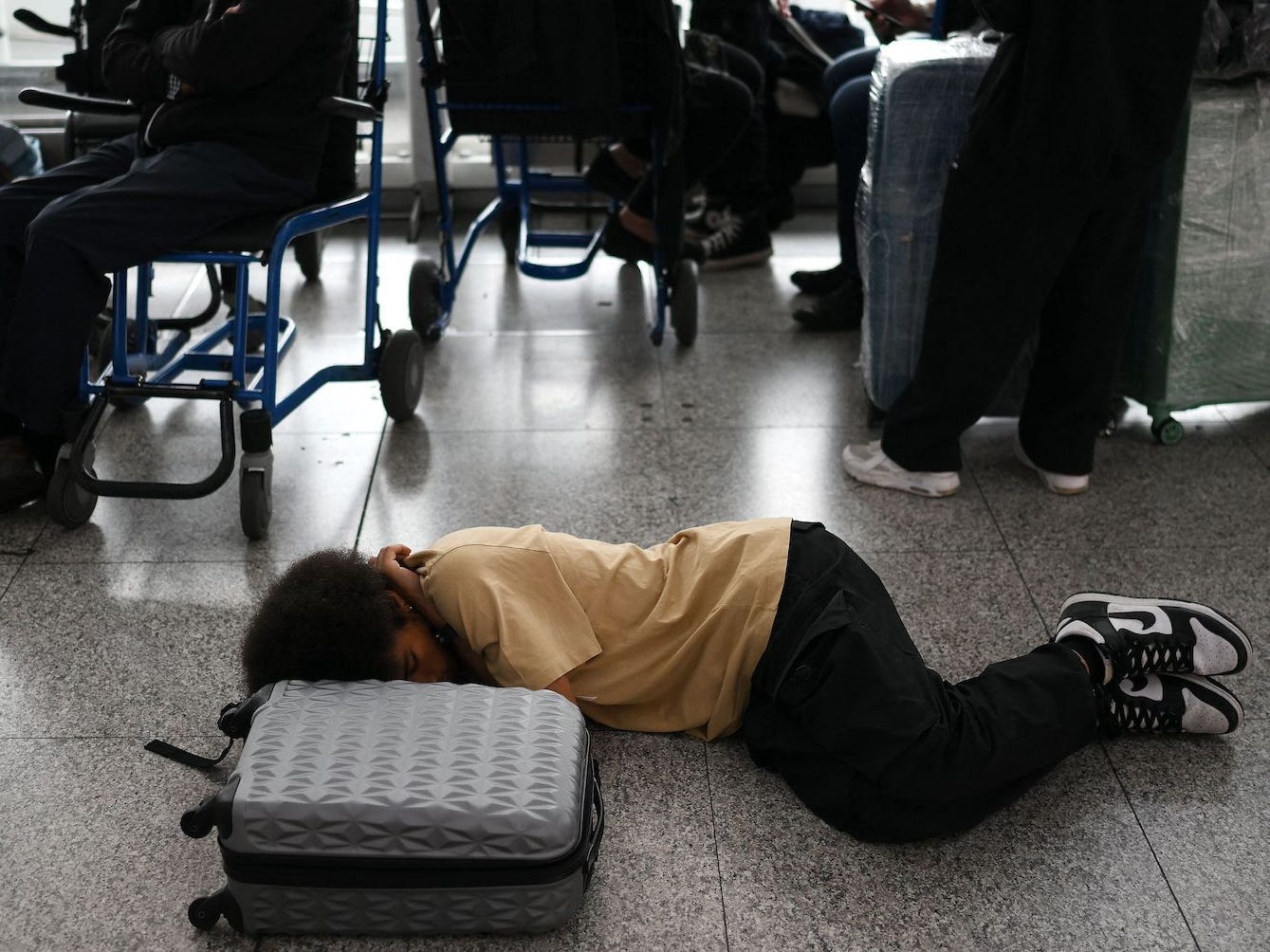 Ein Passagier schläft am Flughafen Stansted neben seinem Gepäck auf dem Boden, während andere Passagiere im Hintergrund sitzen.