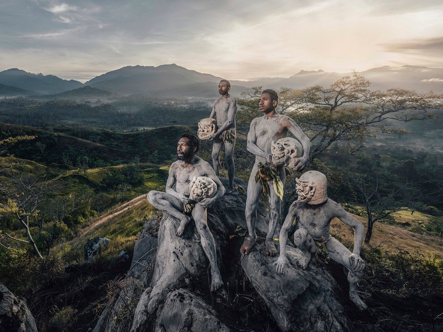 Eine Gruppe von Männern in Körperbemalung sitzt auf einem Felsen und hält Masken vor einer Berglandschaft.