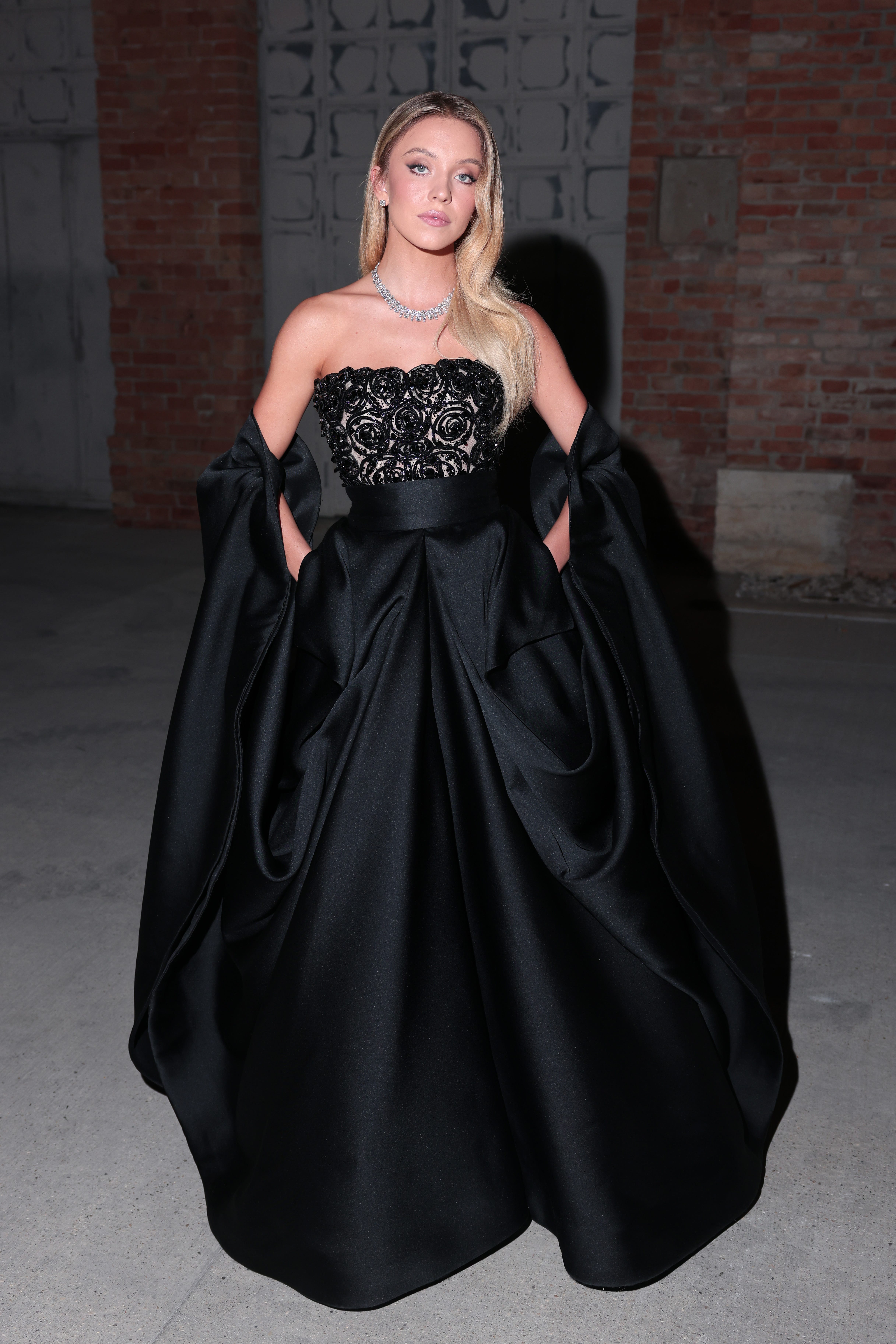 Sydney Sweeney in einem schwarzen trägerlosen Kleid mit Spitzendetails.  Das Kleid hat ein Cape-Detail, das über ihren Ellenbogen sitzt, und sie trägt Diamant-Ohrstecker und eine Diamant-Halskette