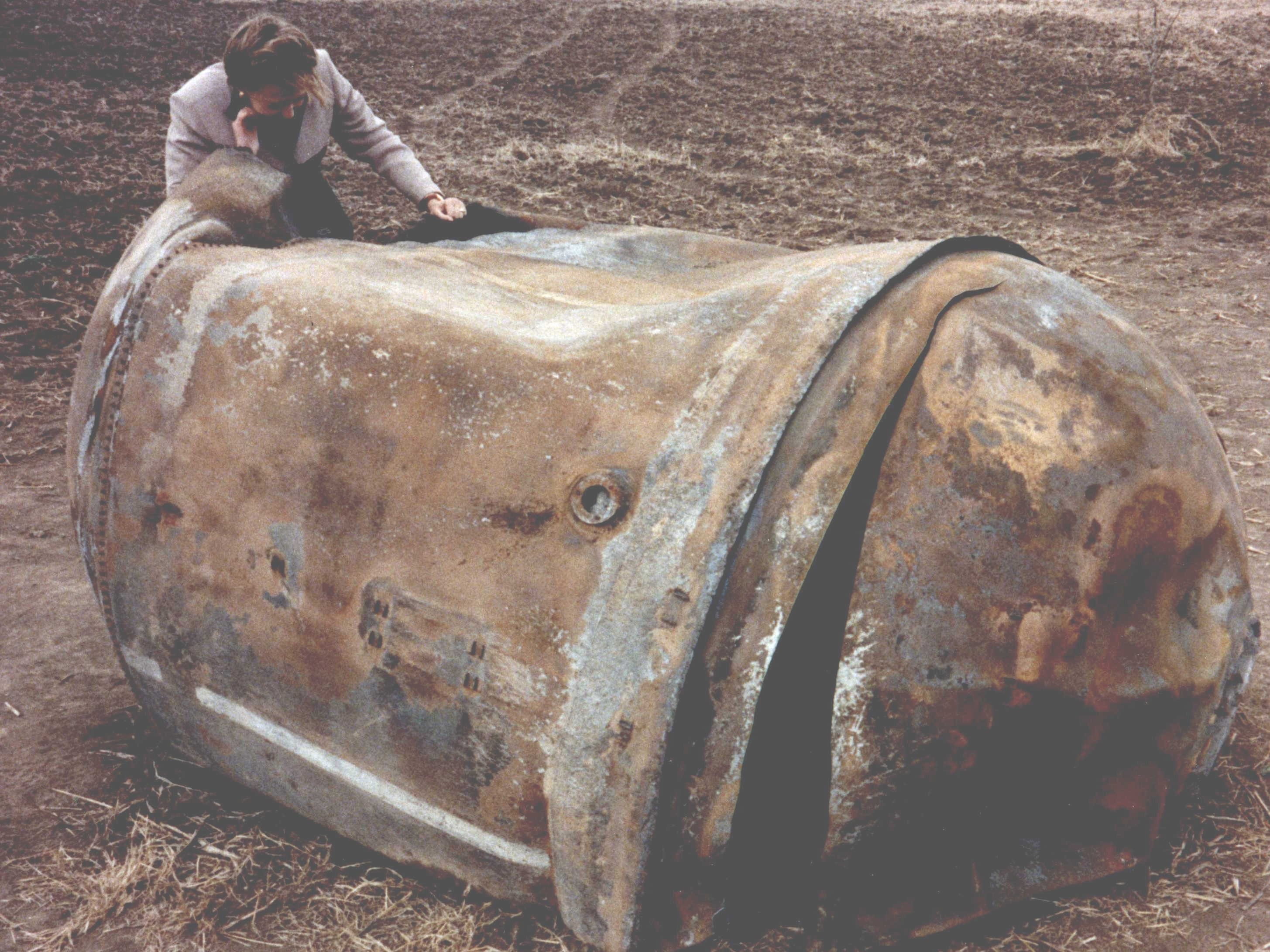 Ein Mann steht über einem Stück Weltraumschrott, das auf einem leeren Feld in Texas landete.  Es sieht aus wie ein riesiger, verrosteter und verbeulter Mülleimer, der auf die Seite gefallen ist.