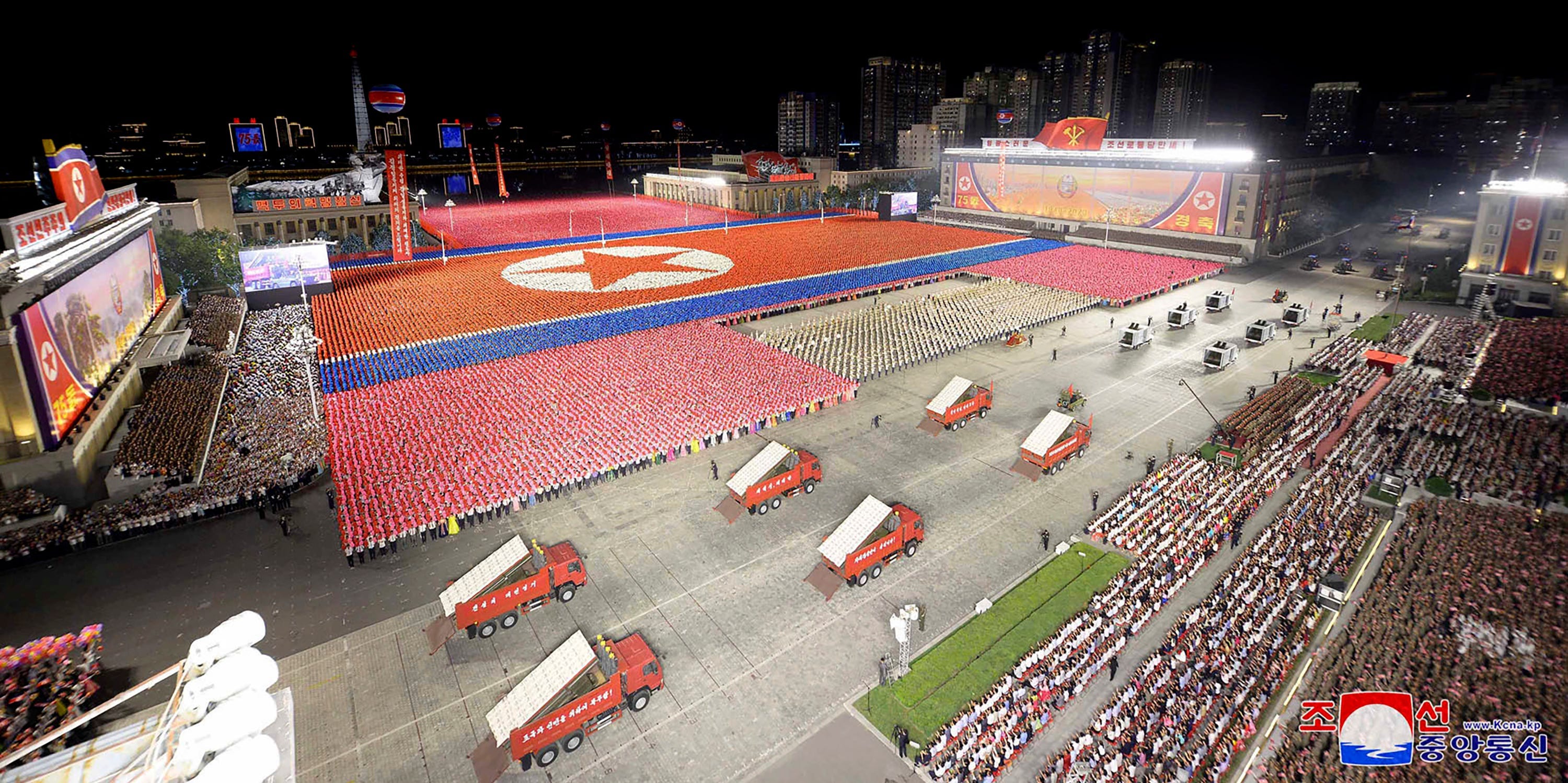 Draufsicht auf einen Platz mit Soldaten in farbigen Uniformen, die die nordkoreanische Flagge formen, während im Vordergrund rote Lastwagen zu sehen sind, aus deren Ladeflächen Raketen auftauchen