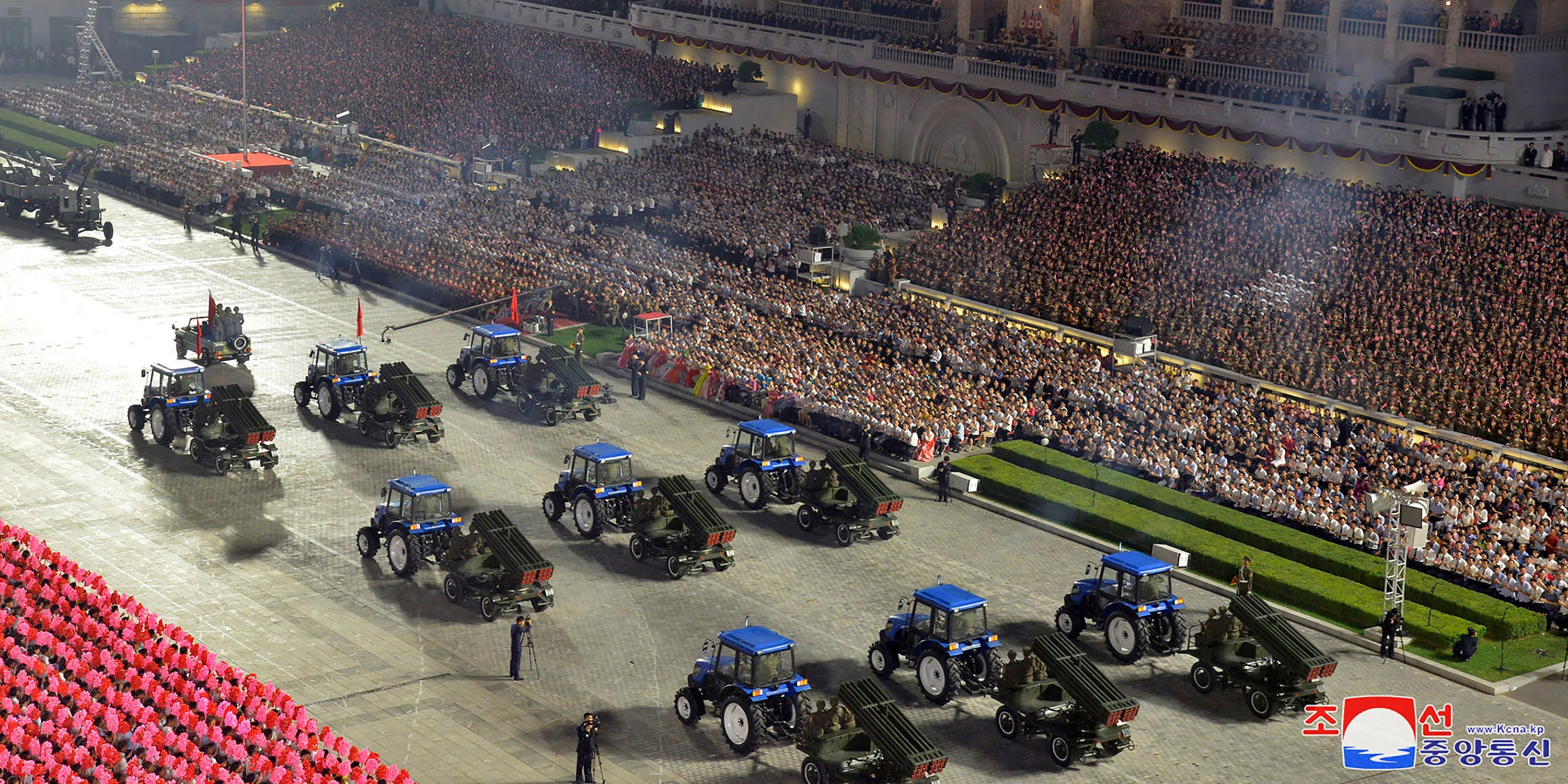 Draufsicht auf neun blaue Traktoren, die Raketenwerfer vor dem Stadion voller Menschen ziehen