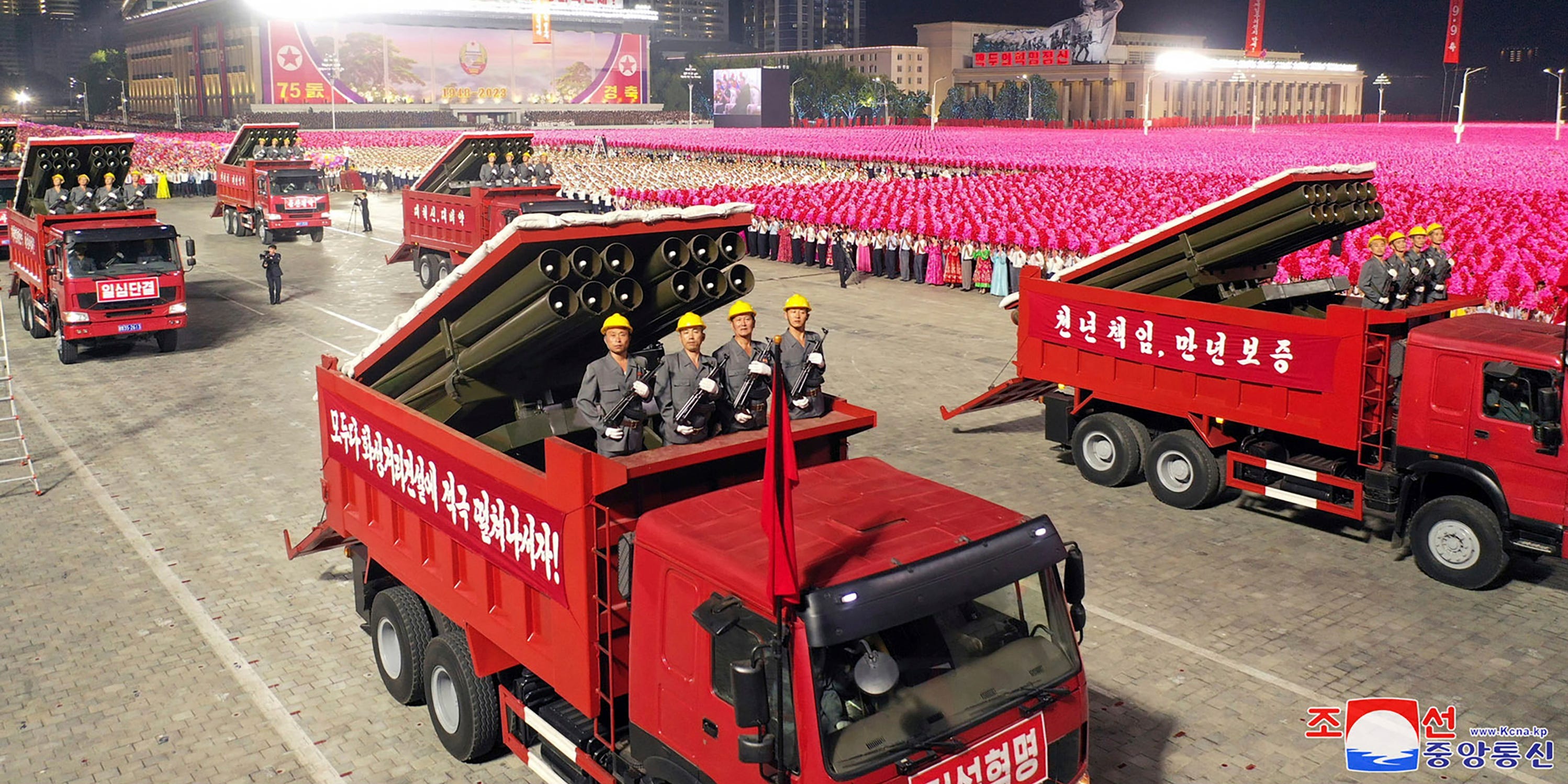 Auf dem roten Lastwagen im Vordergrund tauchen vier Personen in grauen Anzügen und gelben Helmen neben Raketenwerfern auf.  Weitere solcher Lastwagen umgeben es, und in der Ferne stehen Menschenmengen in rosafarbenen Gewändern