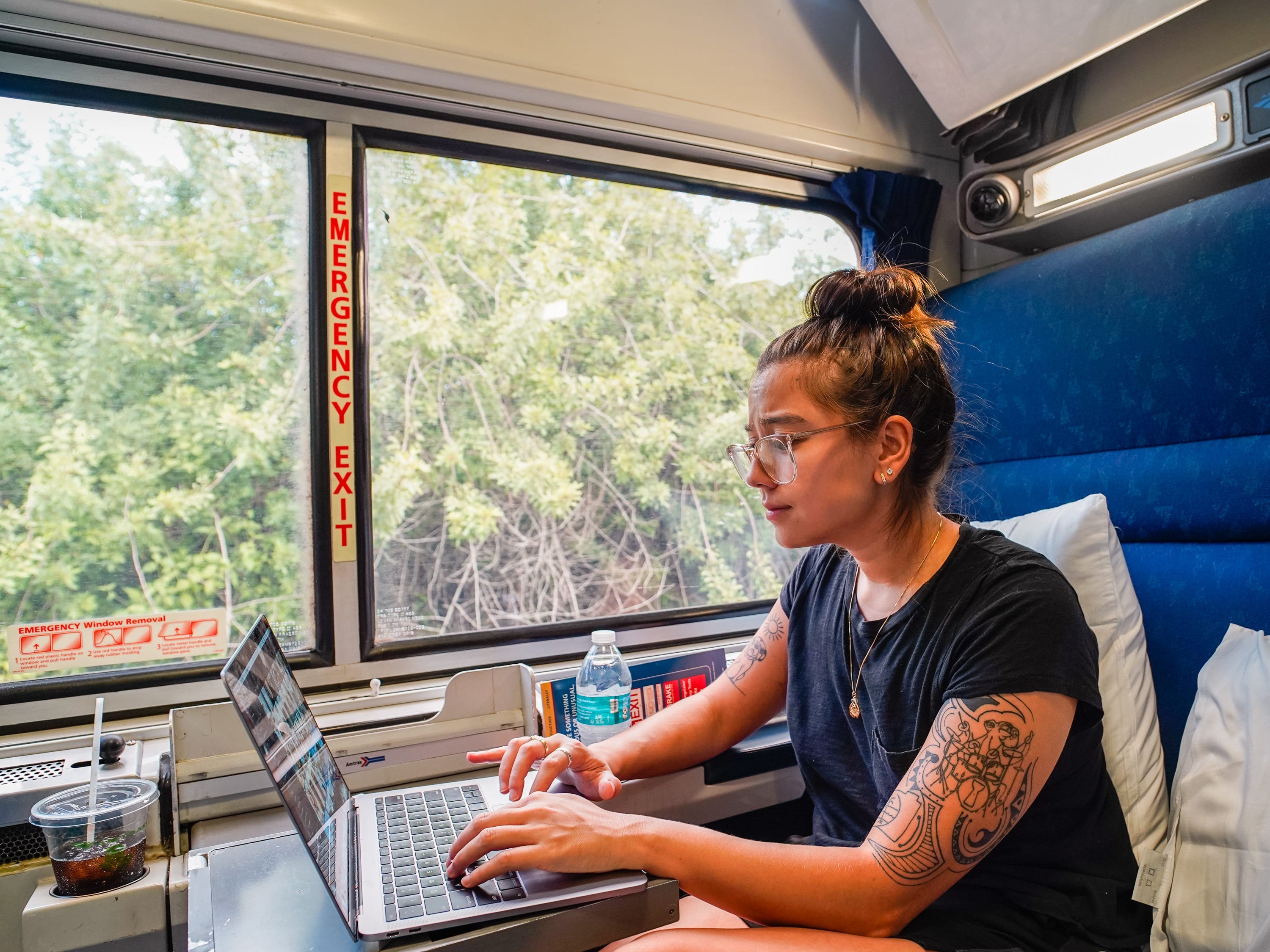 Die Autorin benutzt ihren Laptop, während sie auf einem blauen Sitz sitzt, mit einem Fenster zu ihrer Rechten, durch das man das Grün sehen kann