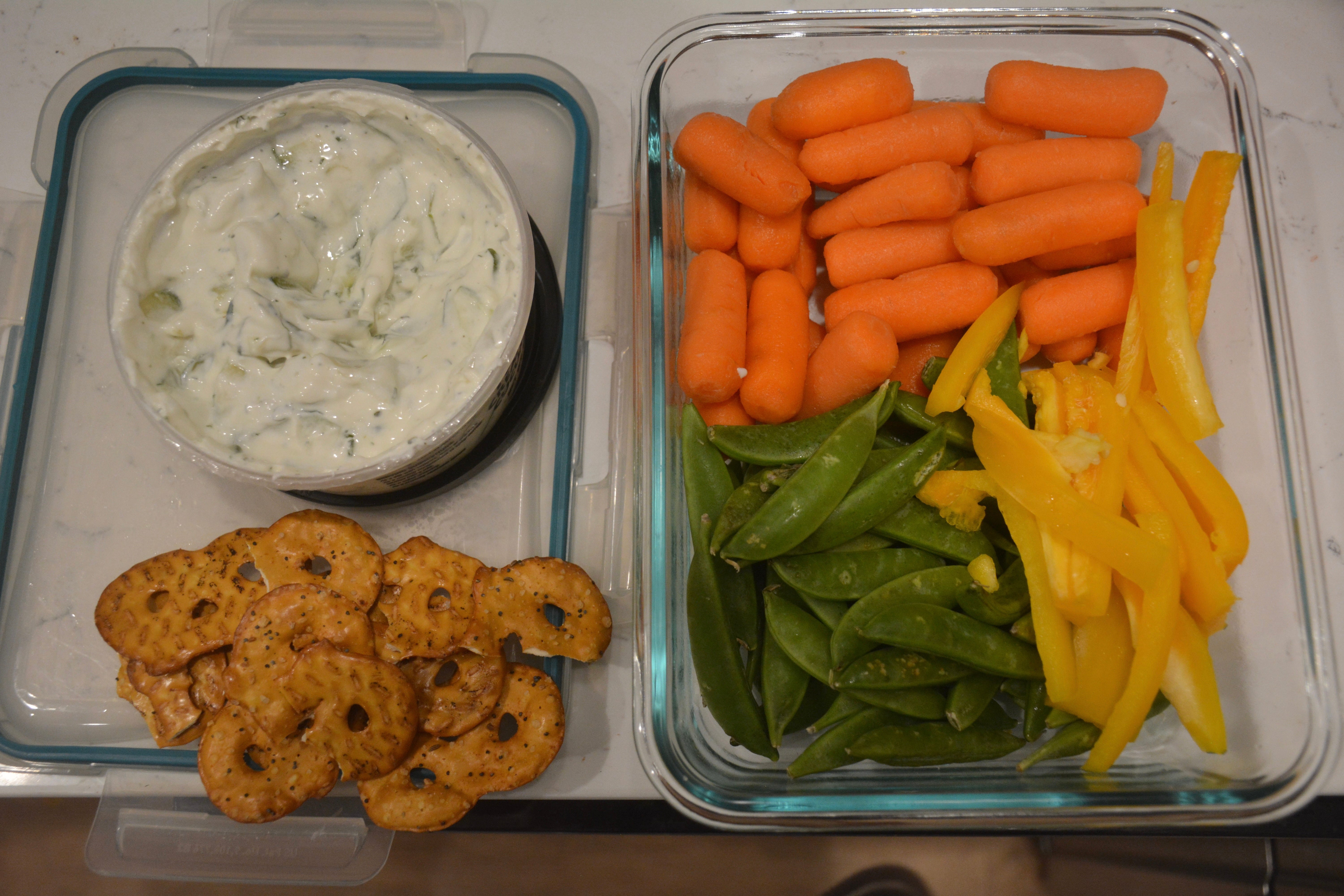 Tupperware mit verzehrfertigem Gemüse, darunter Karotten, Erbsen und gelbe Paprika, daneben Tzatziki und Brezelchips.