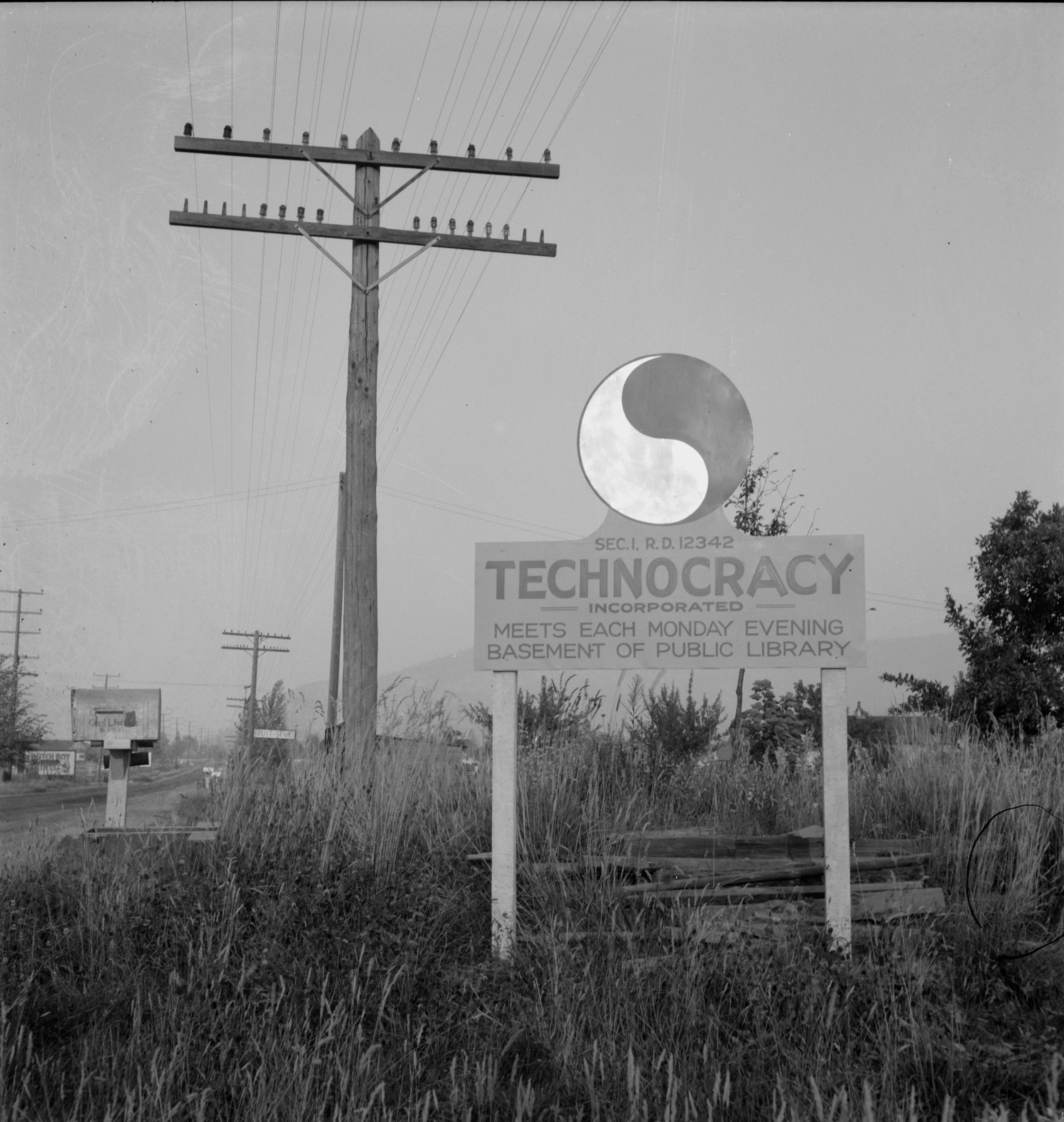 Schwarzweißfoto, Schild mit der Aufschrift „Technocracy Incorporated – trifft sich jeden Montagabend – Keller der öffentlichen Bibliothek“, Rasenfläche, Stromleitungen und leere Straße.