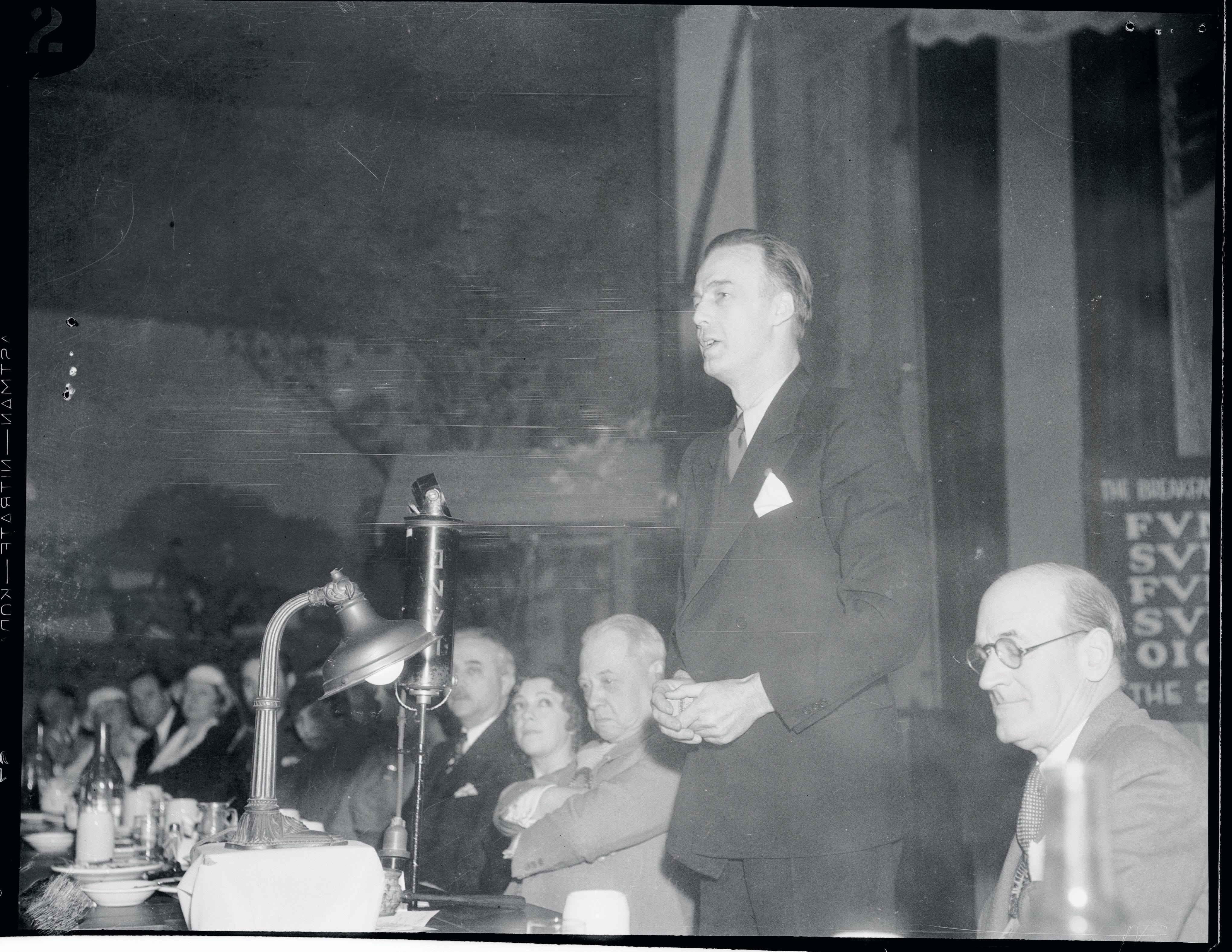 Howard Scott, ein Mann mit nach hinten gekämmtem Haar und Anzug, steht auf und spricht zu einer Menschenmenge, die an einem langen Holztisch sitzt.