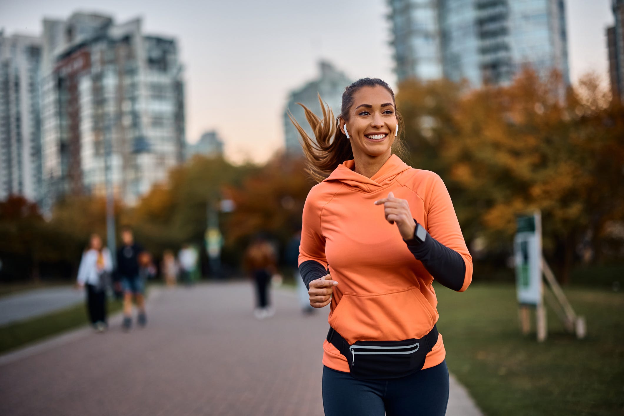 Eine Frau joggt in einem orangefarbenen Sweatshirt und Leggings durch einen Park, trägt Ohrhörer und lächelt