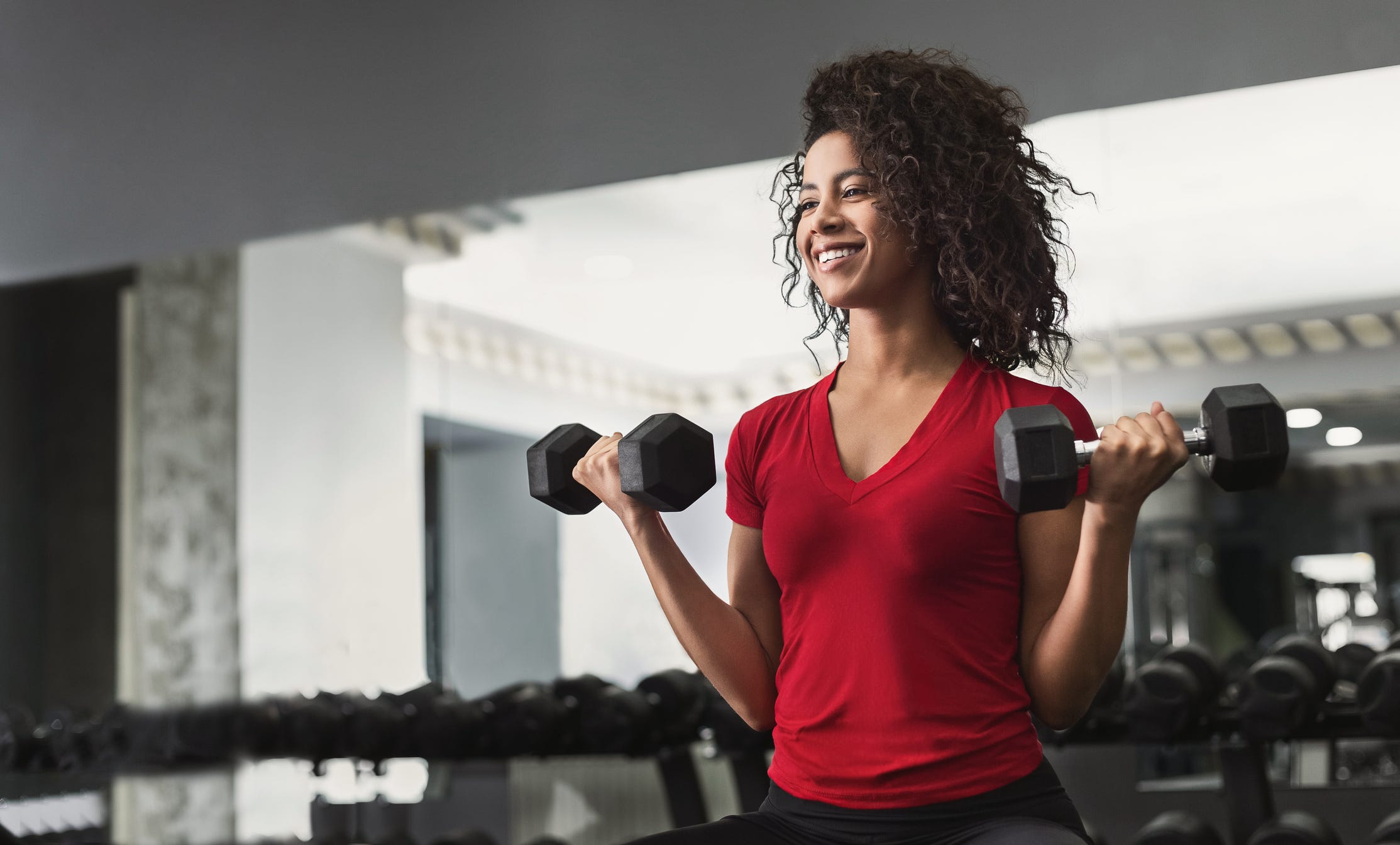 Eine Frau mit lockigem Haar, die in einem Fitnessstudio Hanteln hebt und ein rotes T-Shirt trägt
