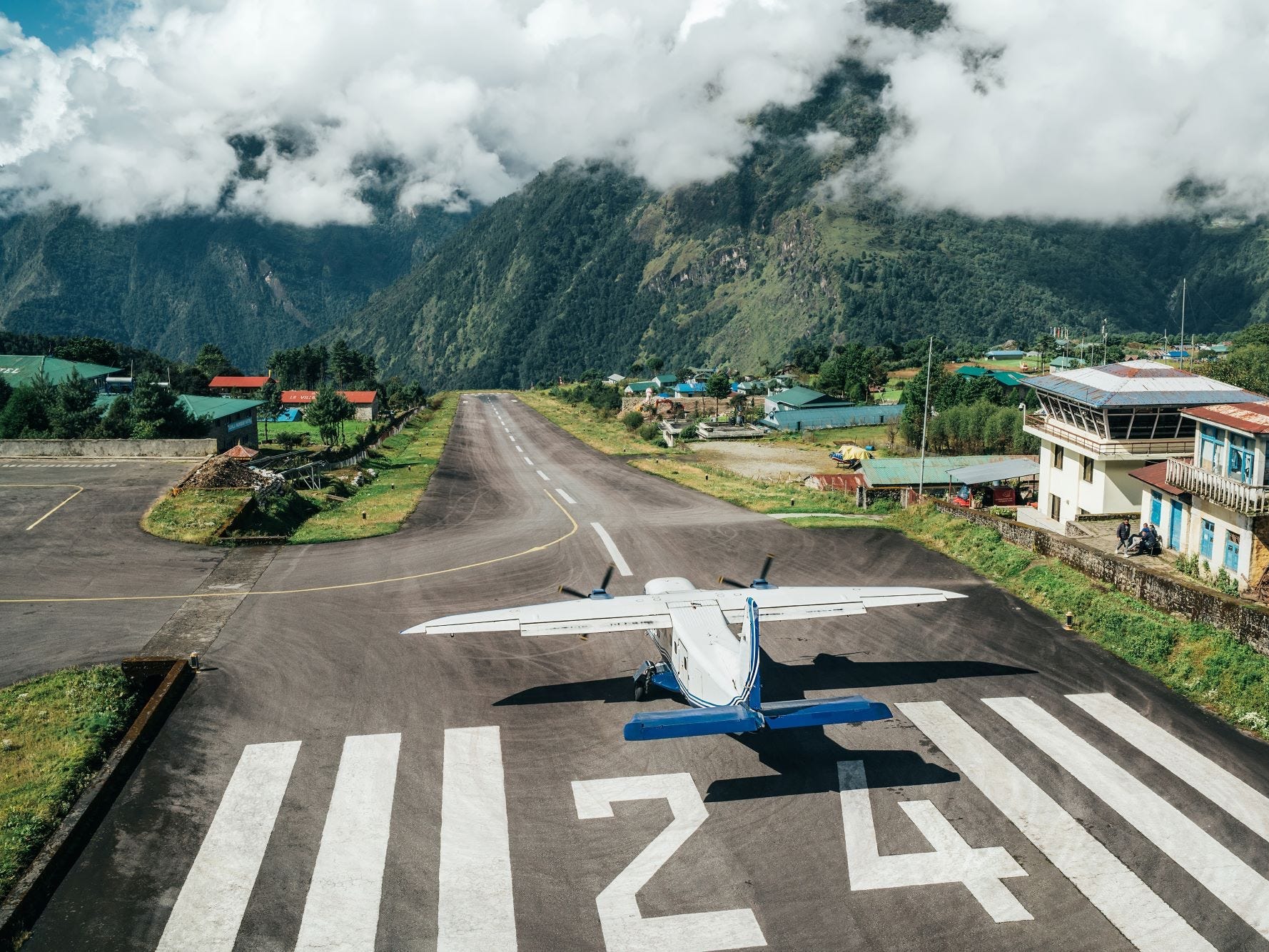 Ein Flugzeug startet von Landebahn 24 am Tenzing-Hillary Airport in Nepal mit Wolken, Bergen und Grün im Hintergrund.