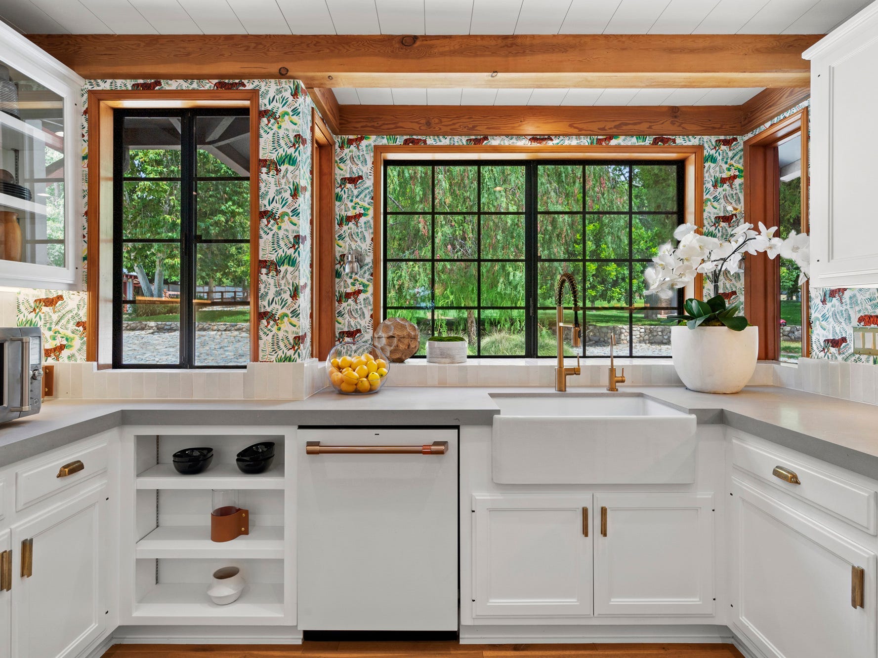 Eine weiße Küchentheke mit mehreren Schubladen und Schränken, einem großen Fenster mit Blick auf einen Baum und weißen Blumen