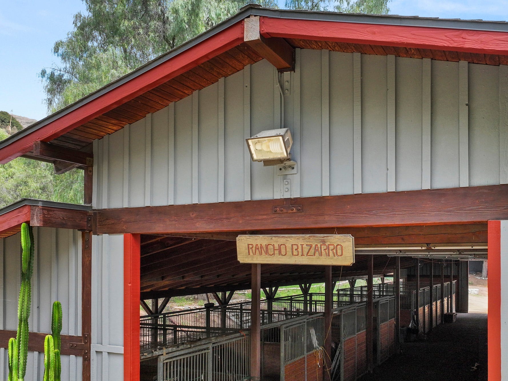 Ein rot-grauer Pferdestall mit offenen Türen und einem Holzschild mit der Aufschrift „Rancho Bizarro“.