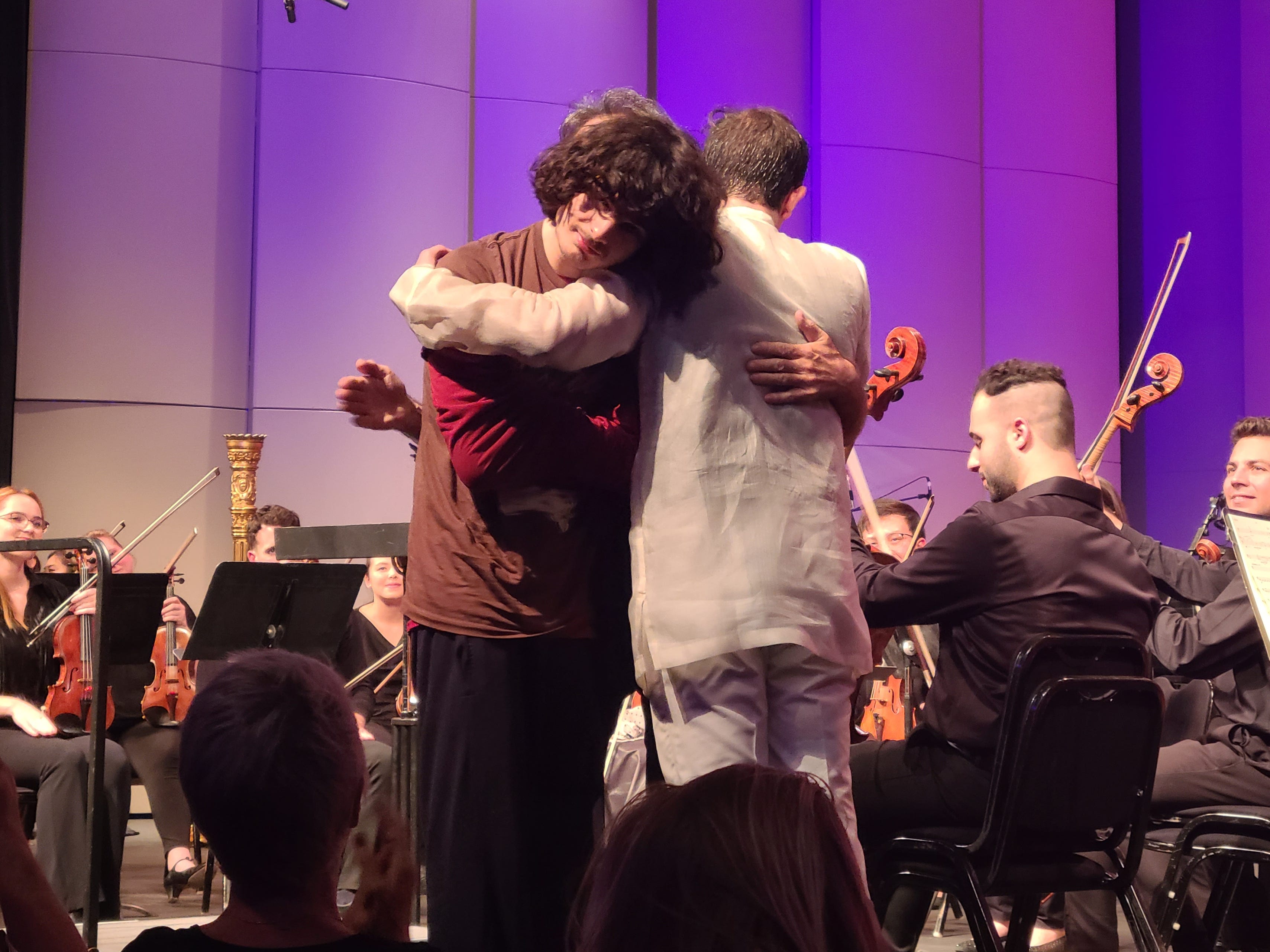 Drei Männer umarmen sich auf der Bühne, hinter ihnen ein Orchester.
