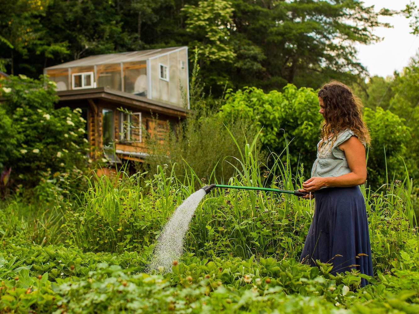 Eine Frau bewässert den Garten mit einem Blockhaus im Hintergrund.