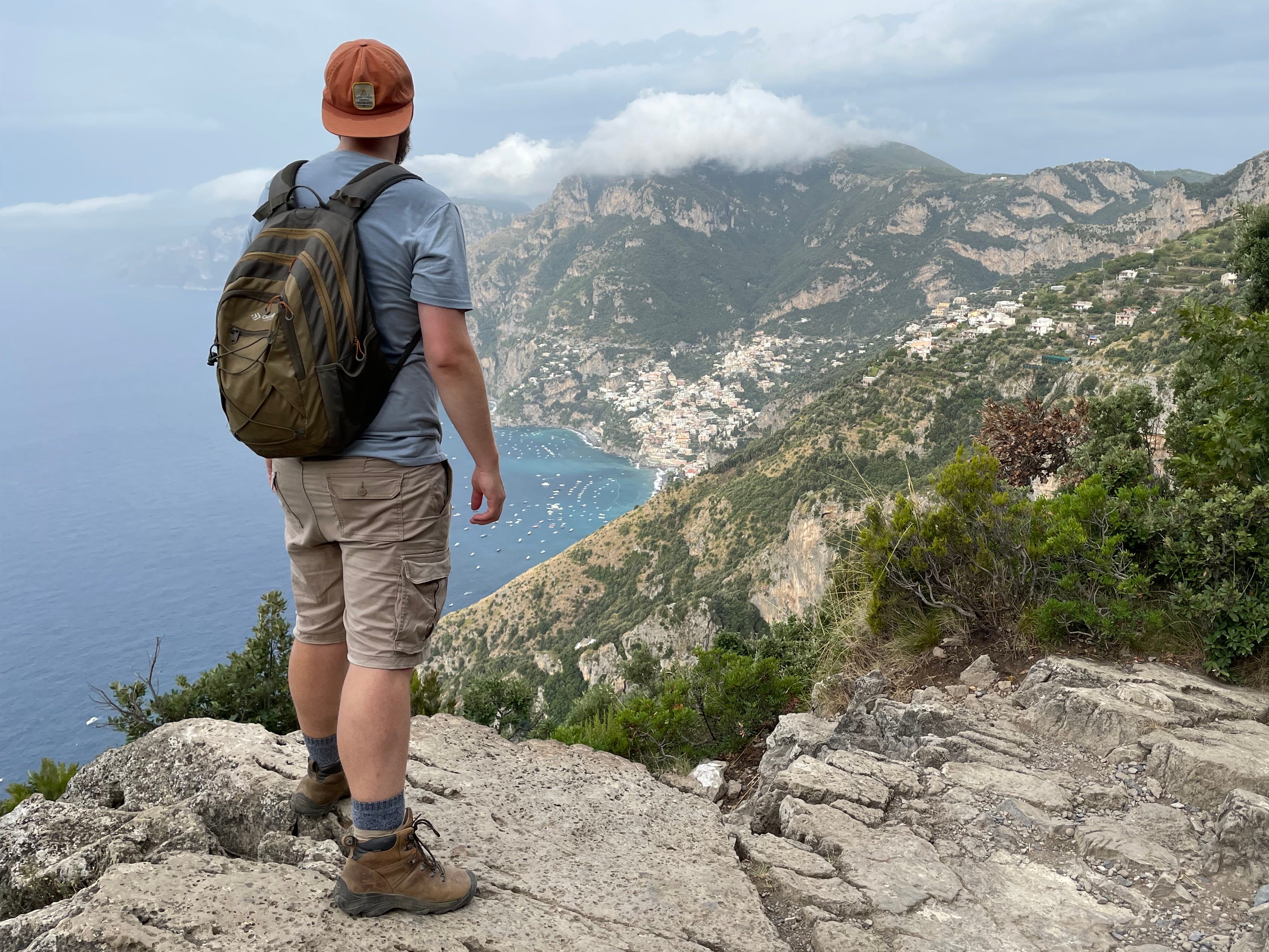 Timothy blickt auf die Aussicht auf einen Berg und trägt einen umgekehrten orangefarbenen Hut, ein blaues Hemd, Khakihosen, einen Rucksack und Wanderschuhe.