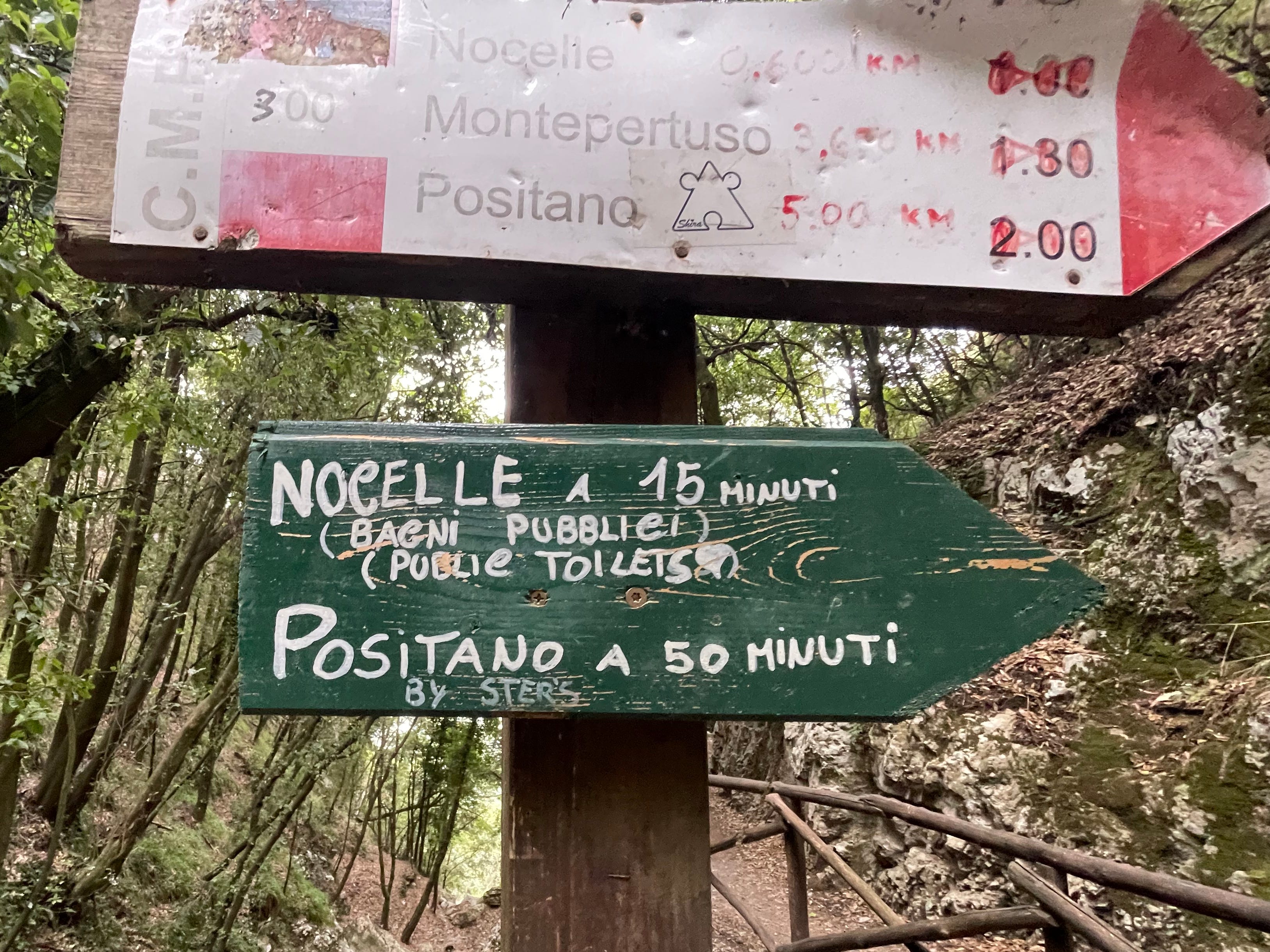 Zwei nach rechts weisende Schilder.  Auf dem unteren Schild steht sowohl auf Italienisch als auch auf Englisch: „Nocelle, 15 Minuten (öffentliche Toiletten)“ und „Positano 50 Minuten über Stufen“.