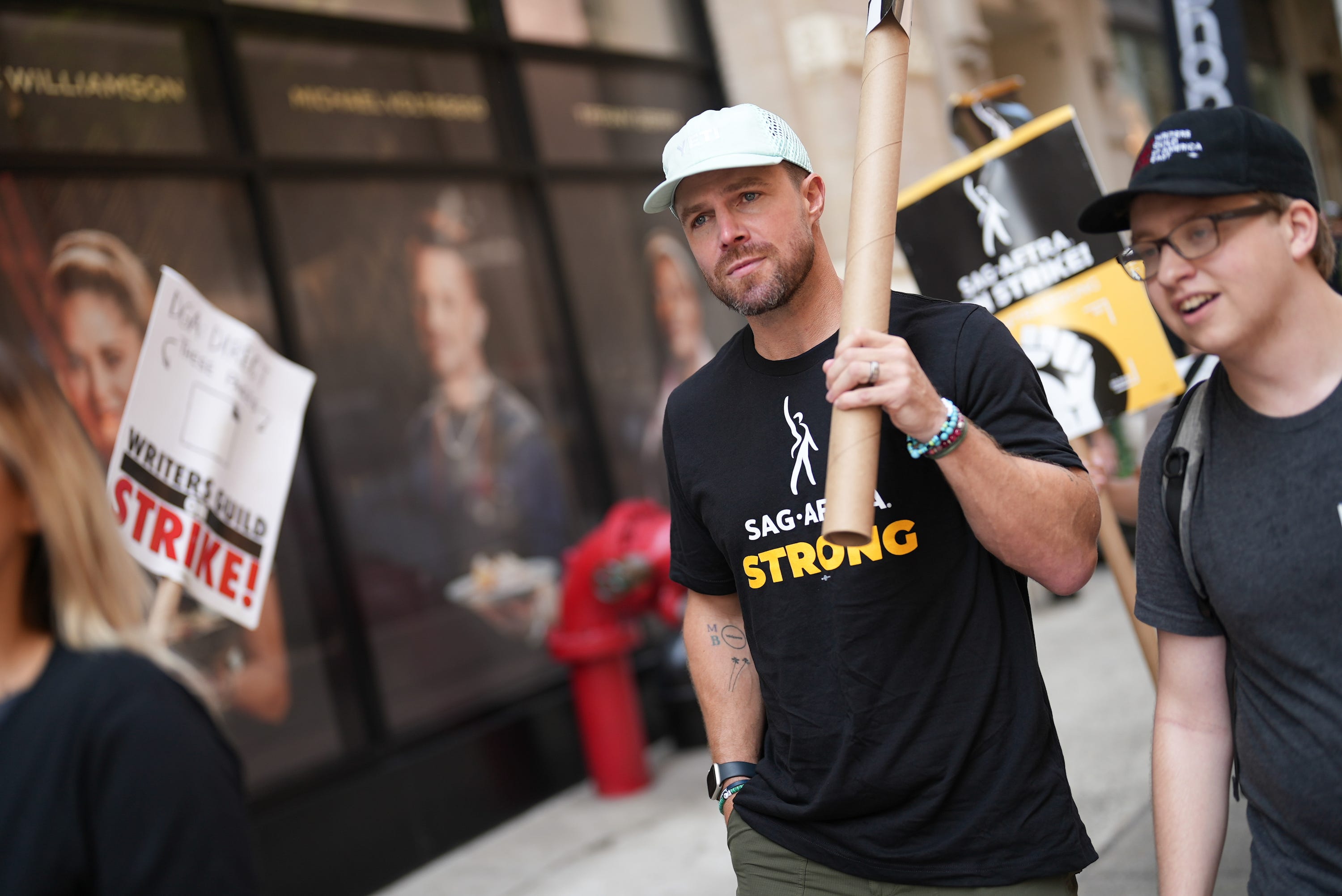 Stephen Amell hält ein Schild in der Hand, während er durch die Streikposten der SAG-AFTRA in New York geht.  Er trägt eine weiße Baseballkappe und ein schwarzes „SAG-AFTRA strong“-T-Shirt.