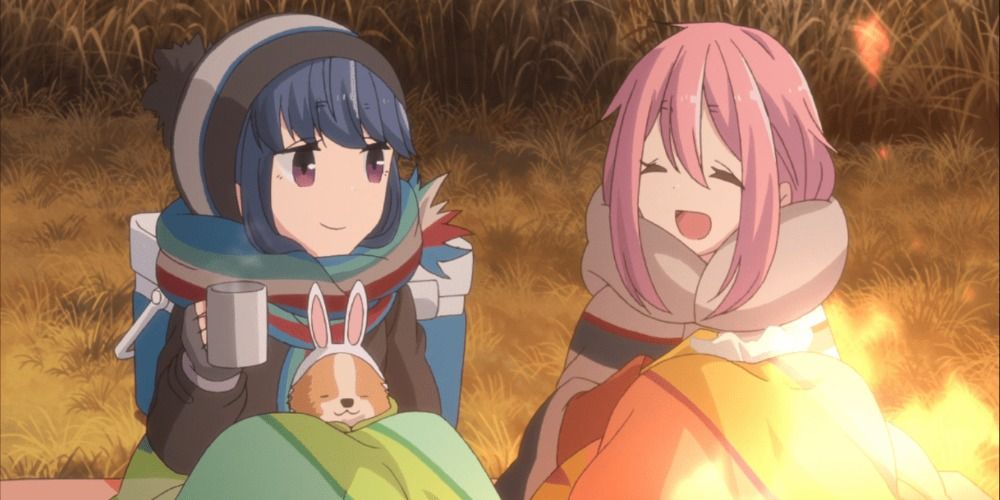 Rin und Nadeshiko vom Laid Back Camp sitzen in Decken gehüllt am Lagerfeuer und lächeln