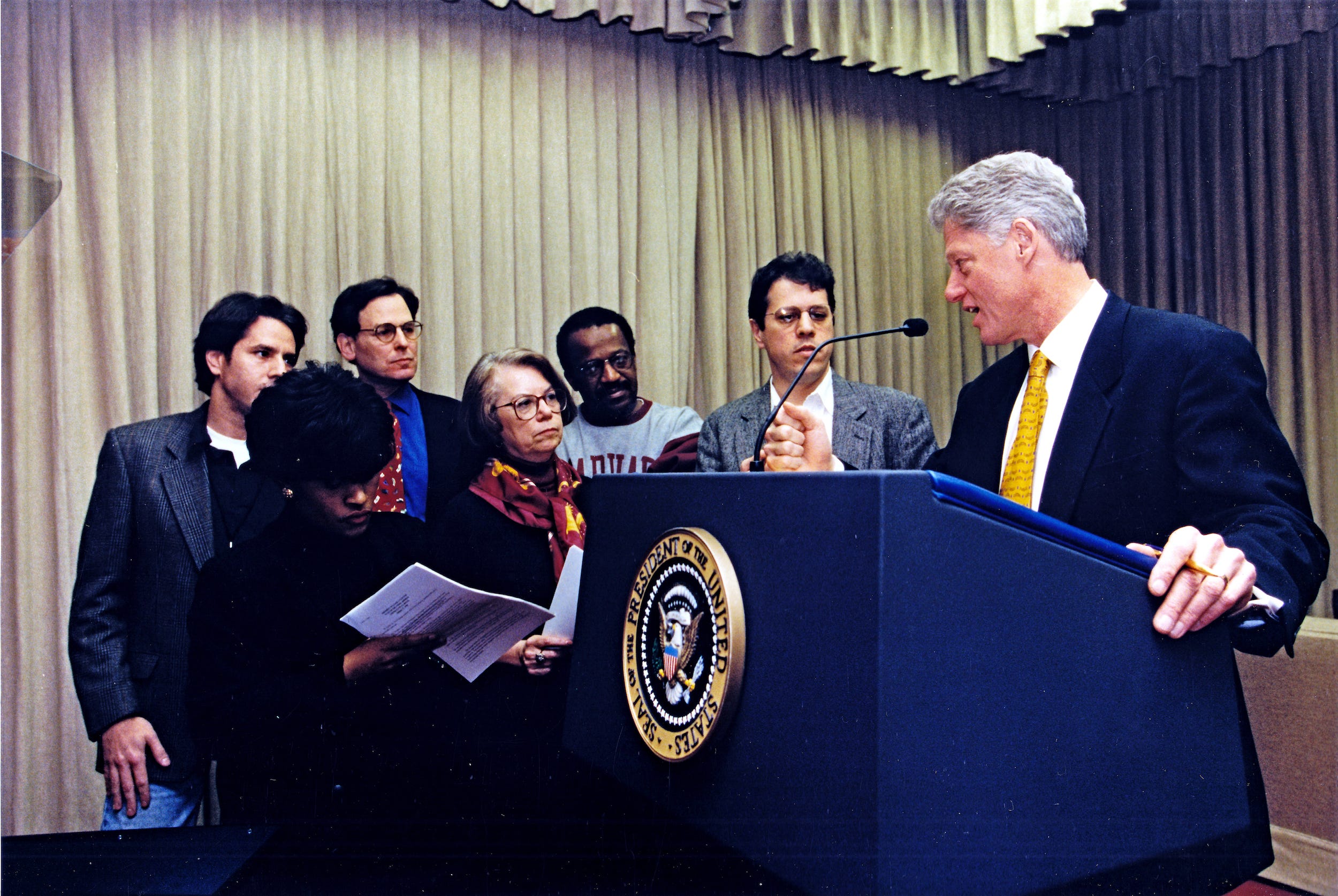 Ein Foto von Bill Clinton auf einem Podium, wie er mit seinen Mitarbeitern spricht.