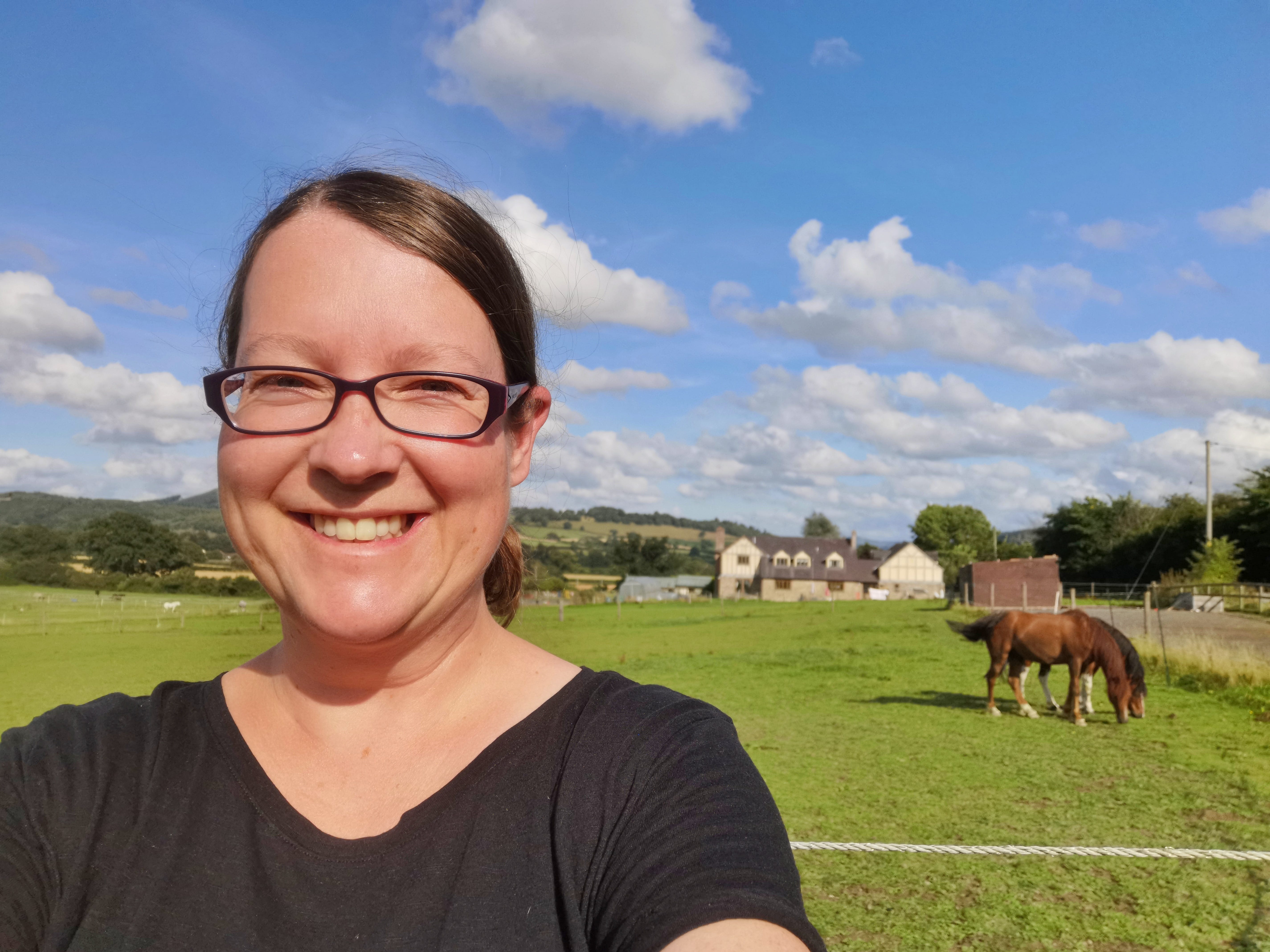 Eine Frau macht ein Selfie vor einer Weide mit grasenden Pferden und einem Bauernhaus im Hintergrund.