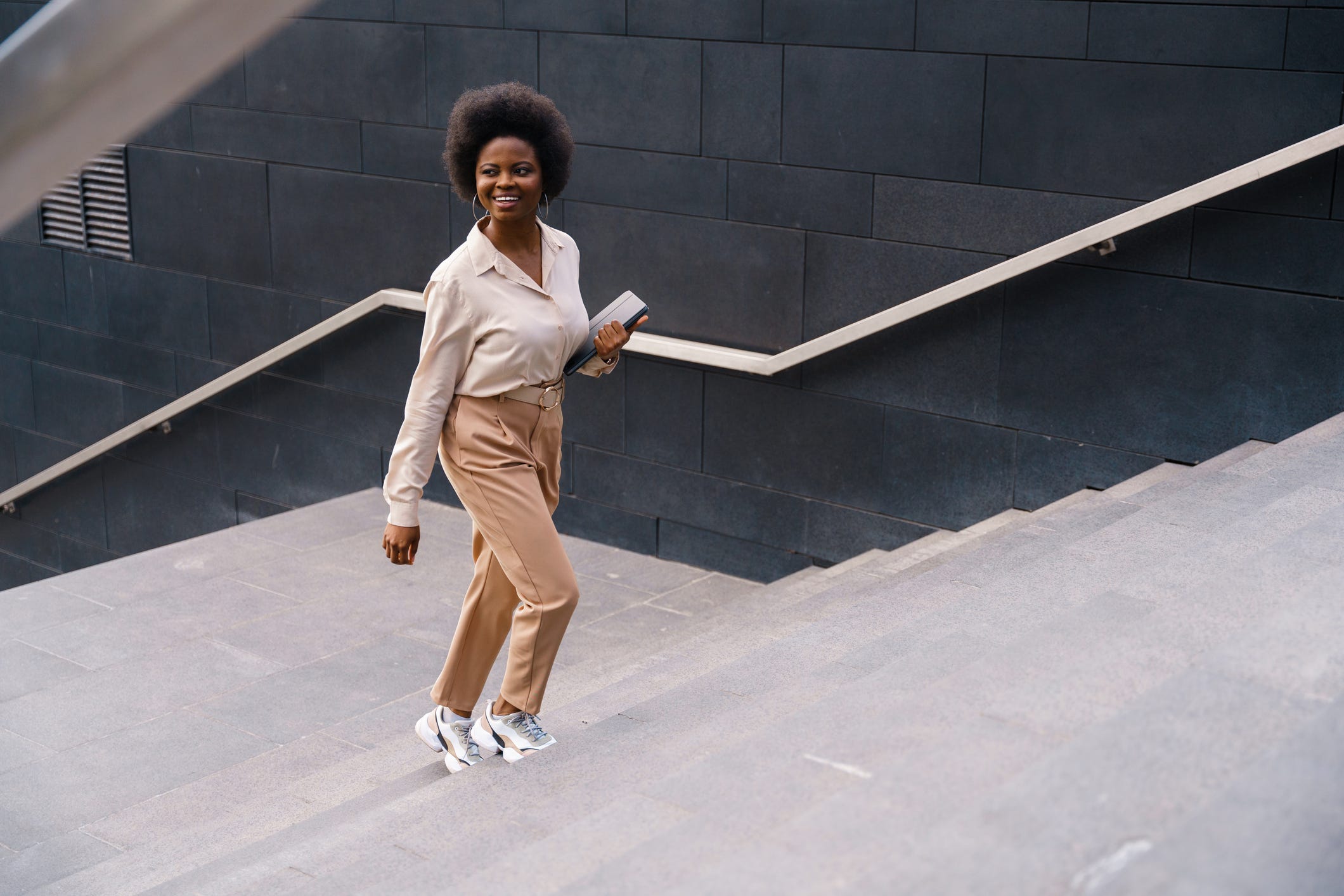 Eine Frau mit natürlicher Frisur, die eine cremefarbene Bluse und eine beige Hose trägt, lächelt und geht eine breite, geflieste Treppe hinauf