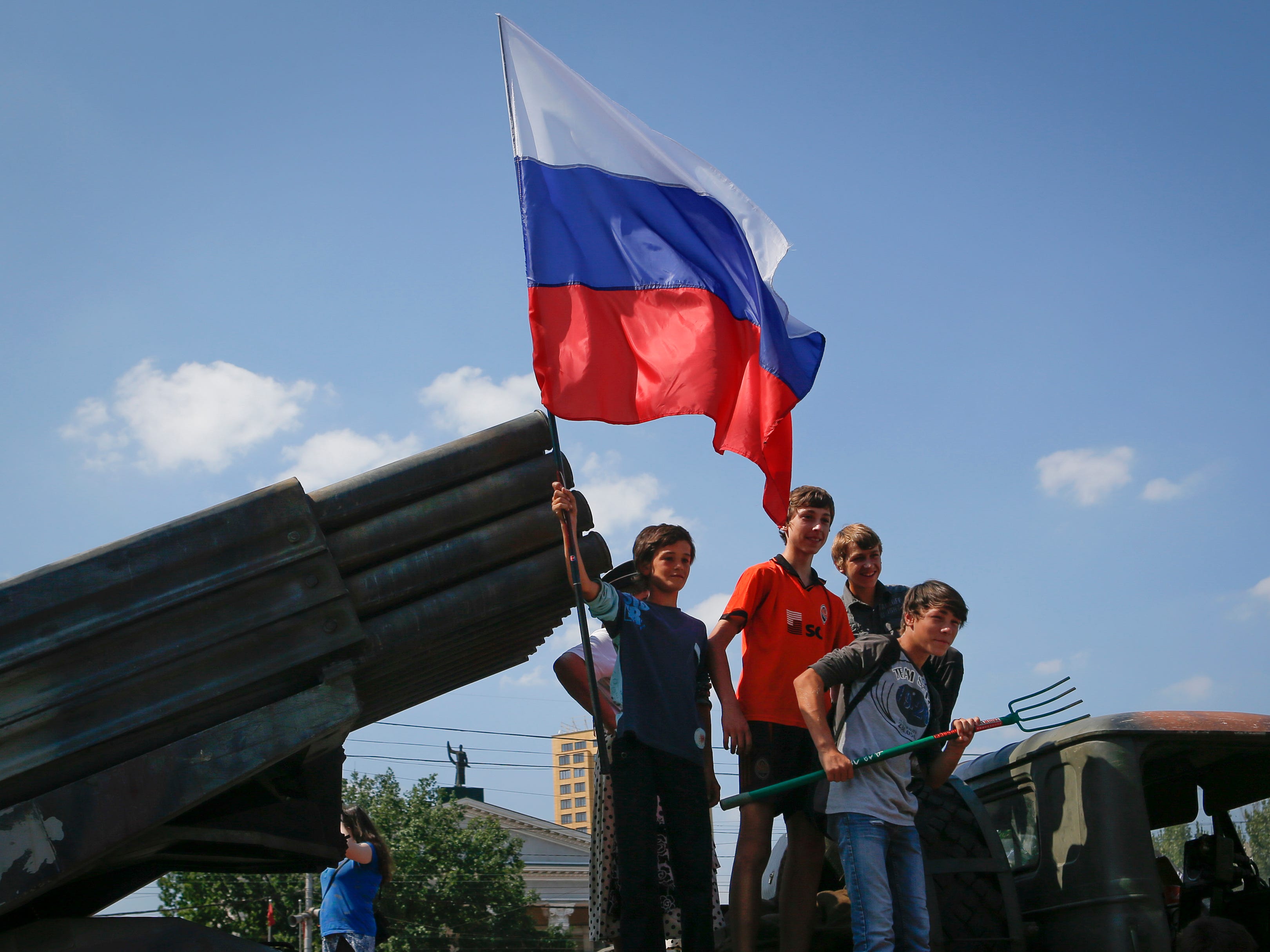 Kinder posieren für ein Foto mit einer russischen Flagge und einer Mistgabel vor einem zerstörten ukrainischen Raketenwerfersystem.