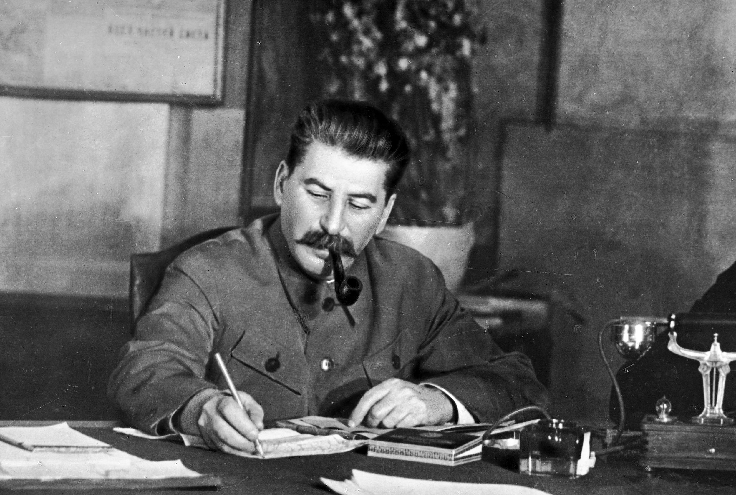 Ein Schwarz-Weiß-Foto von Joseph Stalin, der auf ein Blatt Papier schreibt und an seinem Schreibtisch eine Pfeife raucht.