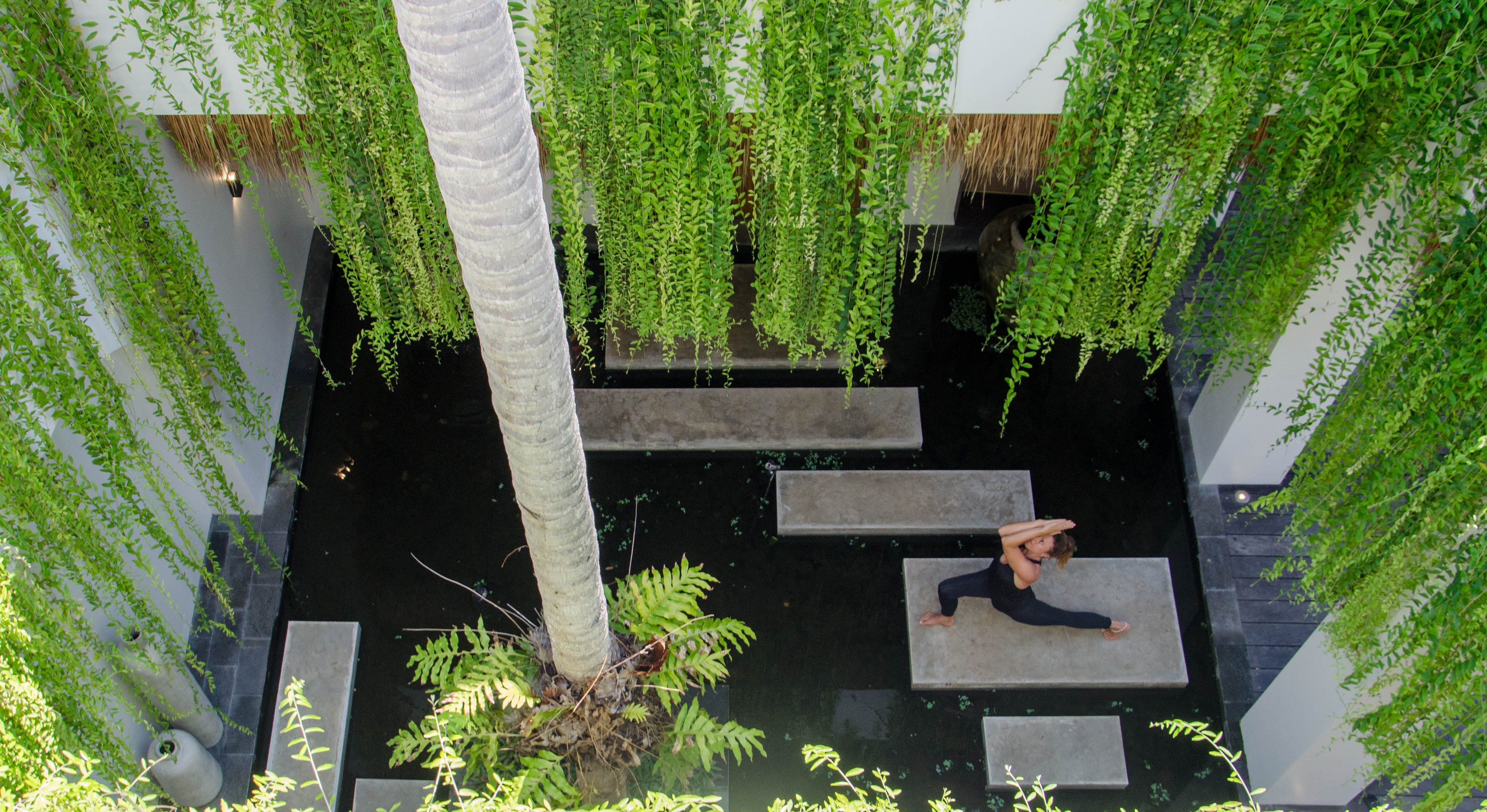 Eine Frau praktiziert Yoga in einem Hotelhof mit Palmen, hängender Vegetation und Wasserspiel