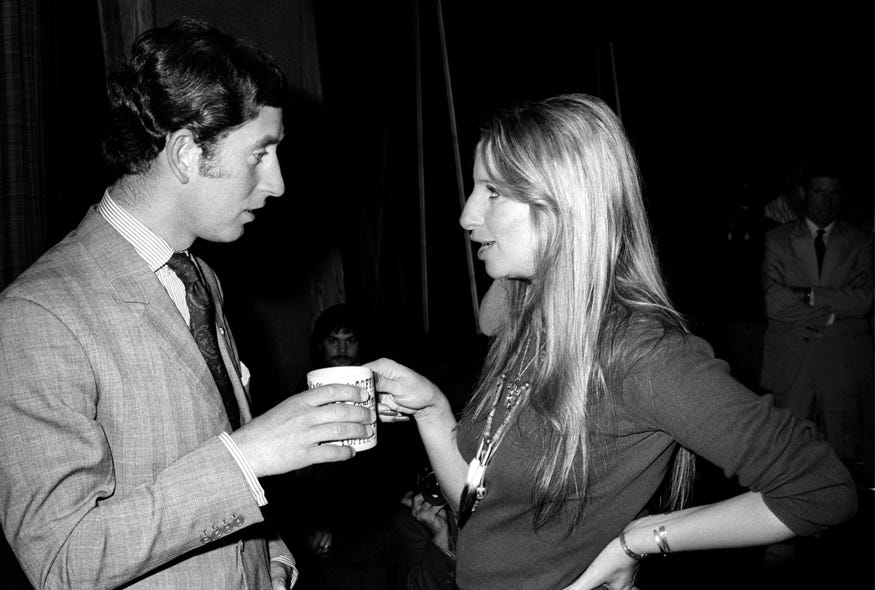 Auf diesem Schwarz-Weiß-Foto aus dem Jahr 1974 steht ein junger König Charles III. in Anzug und Krawatte im Gespräch mit einer jungen Barbra Streisand, die ein langärmeliges Hemd trägt und eine Kaffeetasse in der Hand hält.