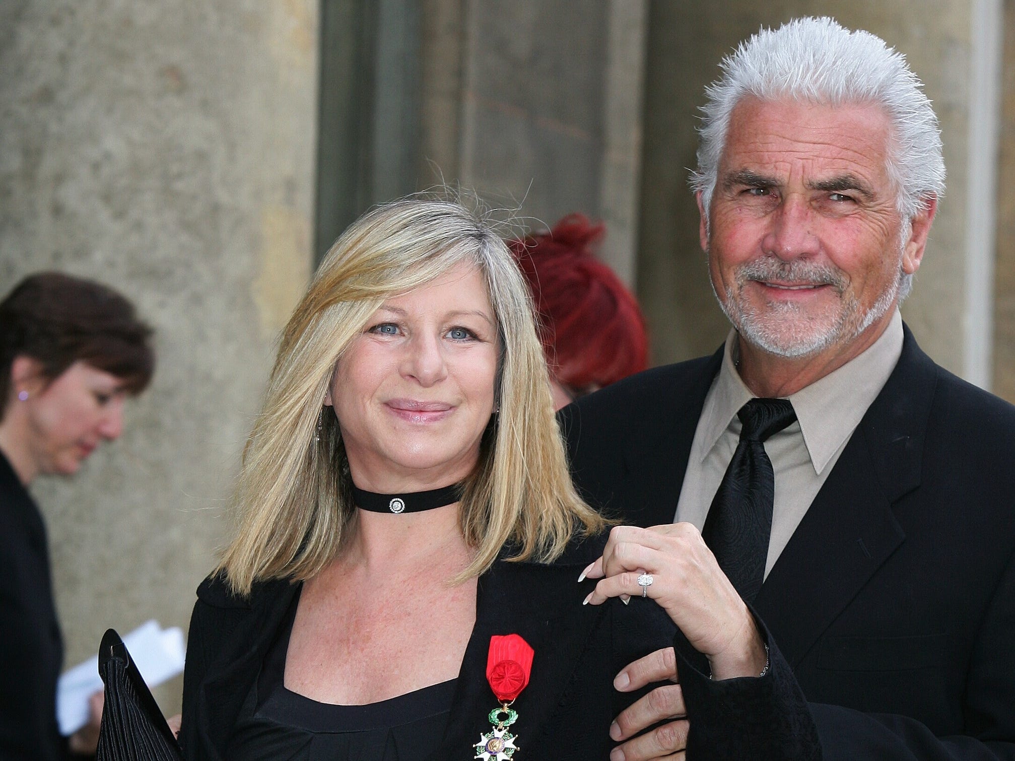 Barbra Streisand, in einem schwarzen Hemd und Blazer, steht mit einer Medaille am Revers.  Ehemann James Brolin steht im schwarzen Anzug hinter ihr und umarmt sie.