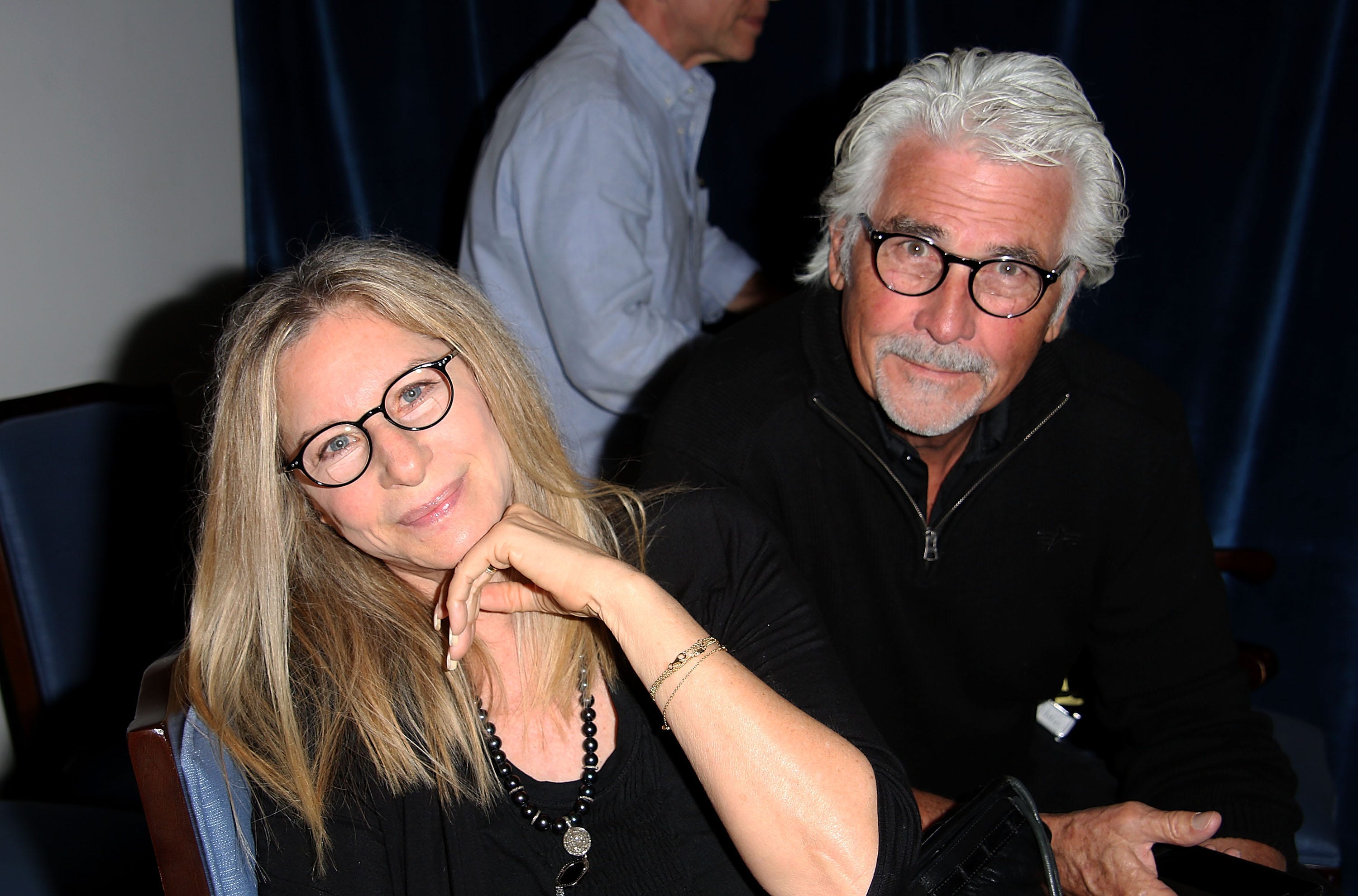 Barbra Streisand, im schwarzen Hemd und mit Brille, lächelt in die Kamera, während Ehemann Jame Brolin, ebenfalls im schwarzen Hemd und mit Brille, hinter ihr sitzt.