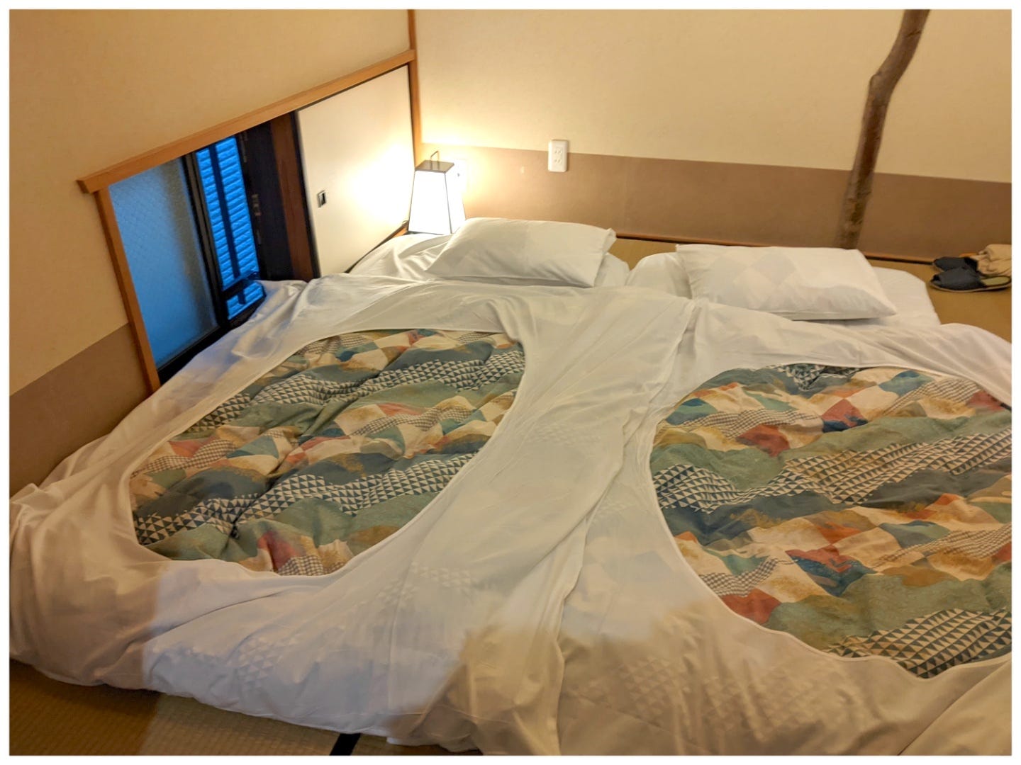 Ein Futonbett im japanischen Stil auf einem Tatami-Mattenboden neben einem kleinen bodentiefen Fenster.  Auf dem Bett liegt ein bunter Bettbezug.