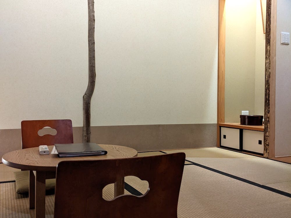 Ein Foto von zwei Holzstühlen ohne Beine, die auf dem Tatami-Mattenboden um einen kleinen Holztisch ruhen.  Gegenüber, in der rechten Ecke des Raumes, befindet sich eine kleine Nische mit einem Regal, in dem sich eine Holzkiste befindet, und darunter zwei Schiebetüren zum Schrank.  Die Wände sind weiß mit einem dünnen, geschnitzten Holzbaum, der von der Decke bis zum Boden reicht.  Die Decke besteht aus Bambusbalken.