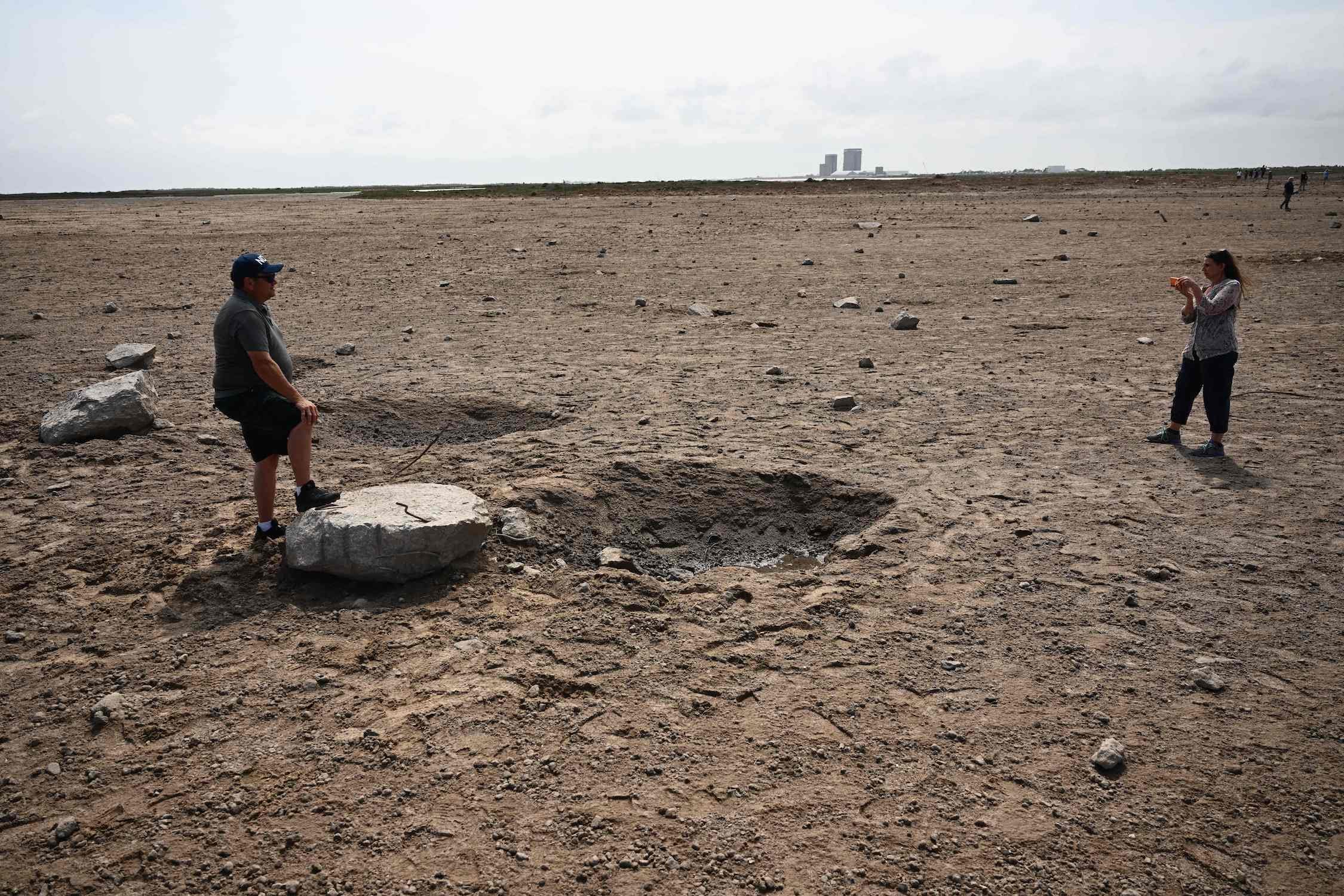 Eine Frau fotografiert einen Mann, der mit einem Fuß auf einem großen Betonbrocken neben einem Krater in einem trockenen Erdfeld steht