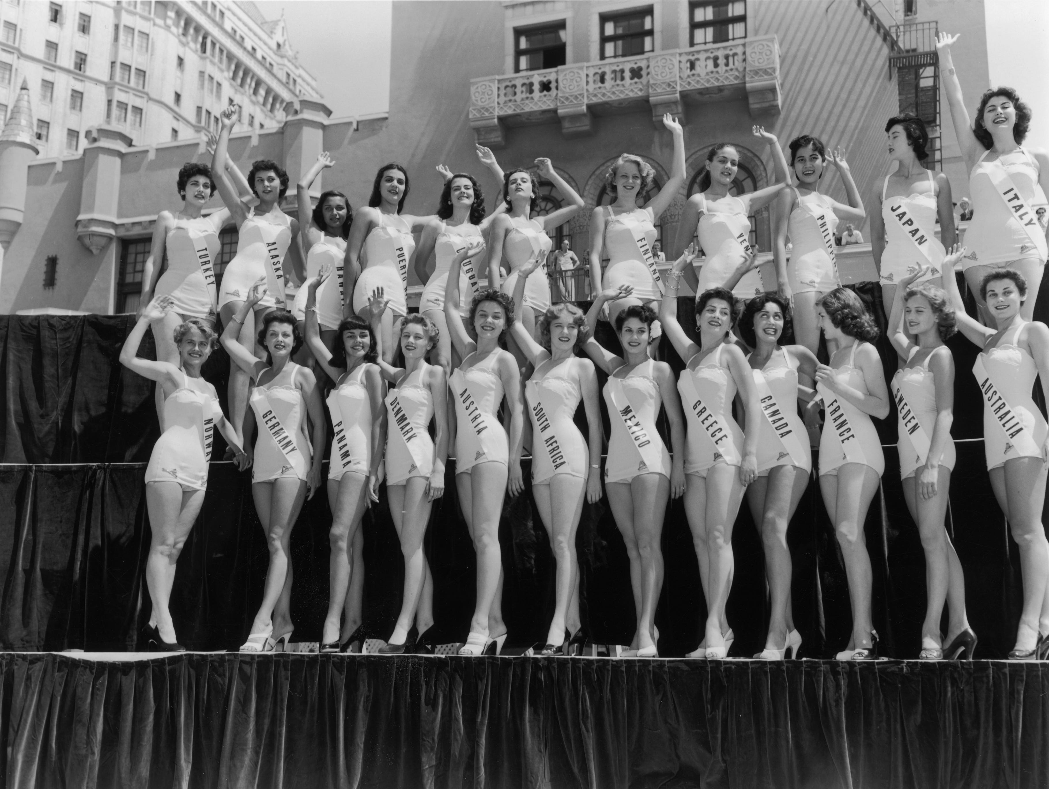 um 1953: Gruppenporträt in voller Länge von Miss Universe-Kandidaten, die winken, während sie in zwei Reihen auf einer Plattform in Long Beach, Kalifornien, stehen.  Alle tragen Badeanzüge und Schuhe mit hohen Absätzen.