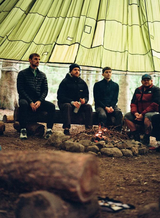 Vier Männer sitzen auf einem Baumstamm unter einem Baldachin, im Vordergrund ein Lagerfeuer