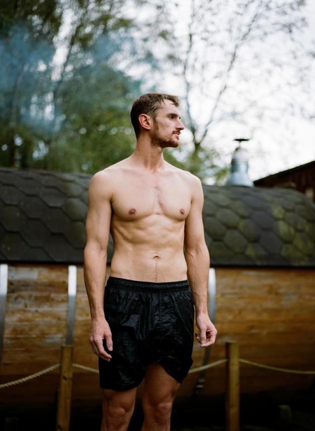 Ein Mann mit nacktem Oberkörper und schwarzen Shorts steht vor einer Sauna