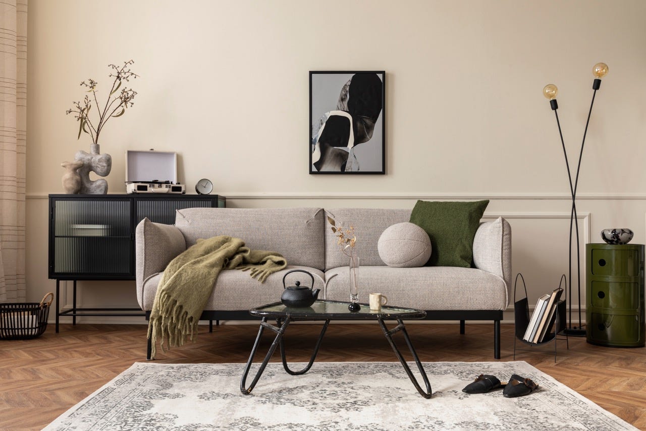 Graue Couch vor schwarzem Metall- und Glastisch vor beigefarbener Wand mit Kunstwerken