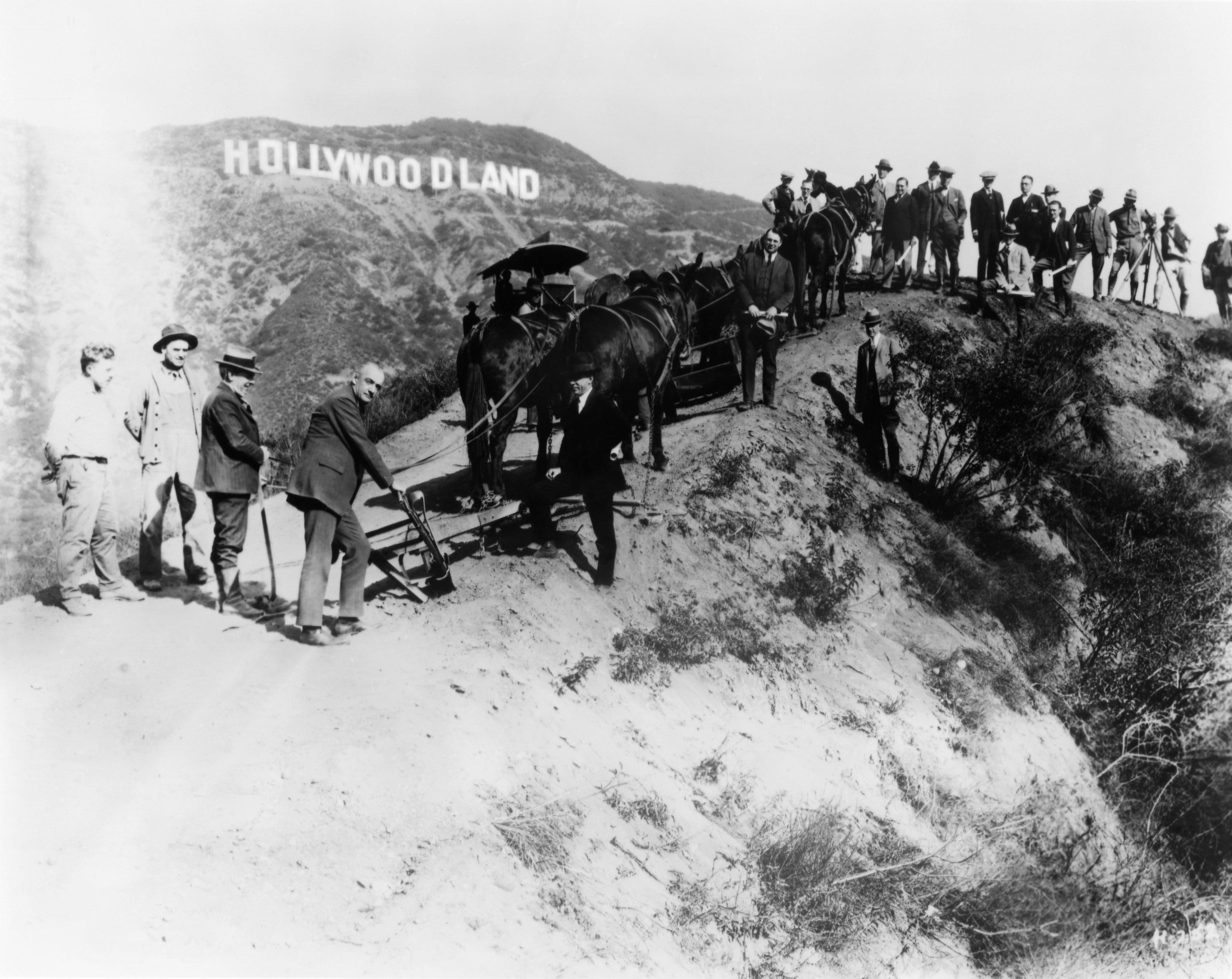Eine Gruppe von Männern, vermutlich Vermessungsingenieure und Bauarbeiter, die an der neuen Wohnsiedlung Hollywoodland arbeiten, posieren für ein Porträt unter dem Schild, das etwa 1925 in Los Angeles, Kalifornien, zur Werbung für das Gelände aufgestellt wurde.