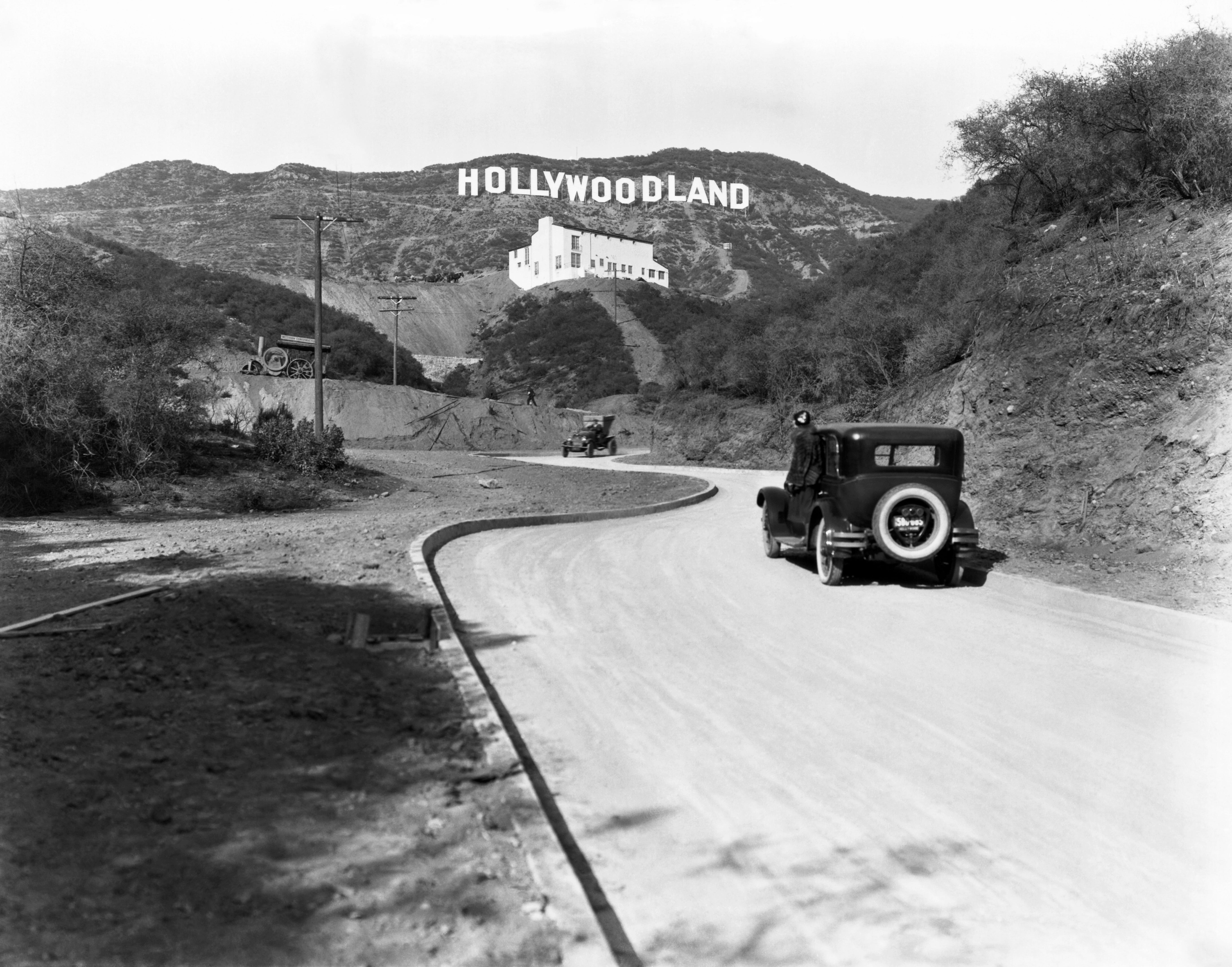 Ein Schild wirbt für die Eröffnung der Wohnsiedlung Hollywoodland in den Hügeln am Mulholland Drive mit Blick auf Los Angeles, Hollywood, Los Angeles, Kalifornien, um 1924. Das weiße Gebäude unter dem Schild ist die Kanst Art Gallery, die am 1. April 1924 eröffnet wurde.