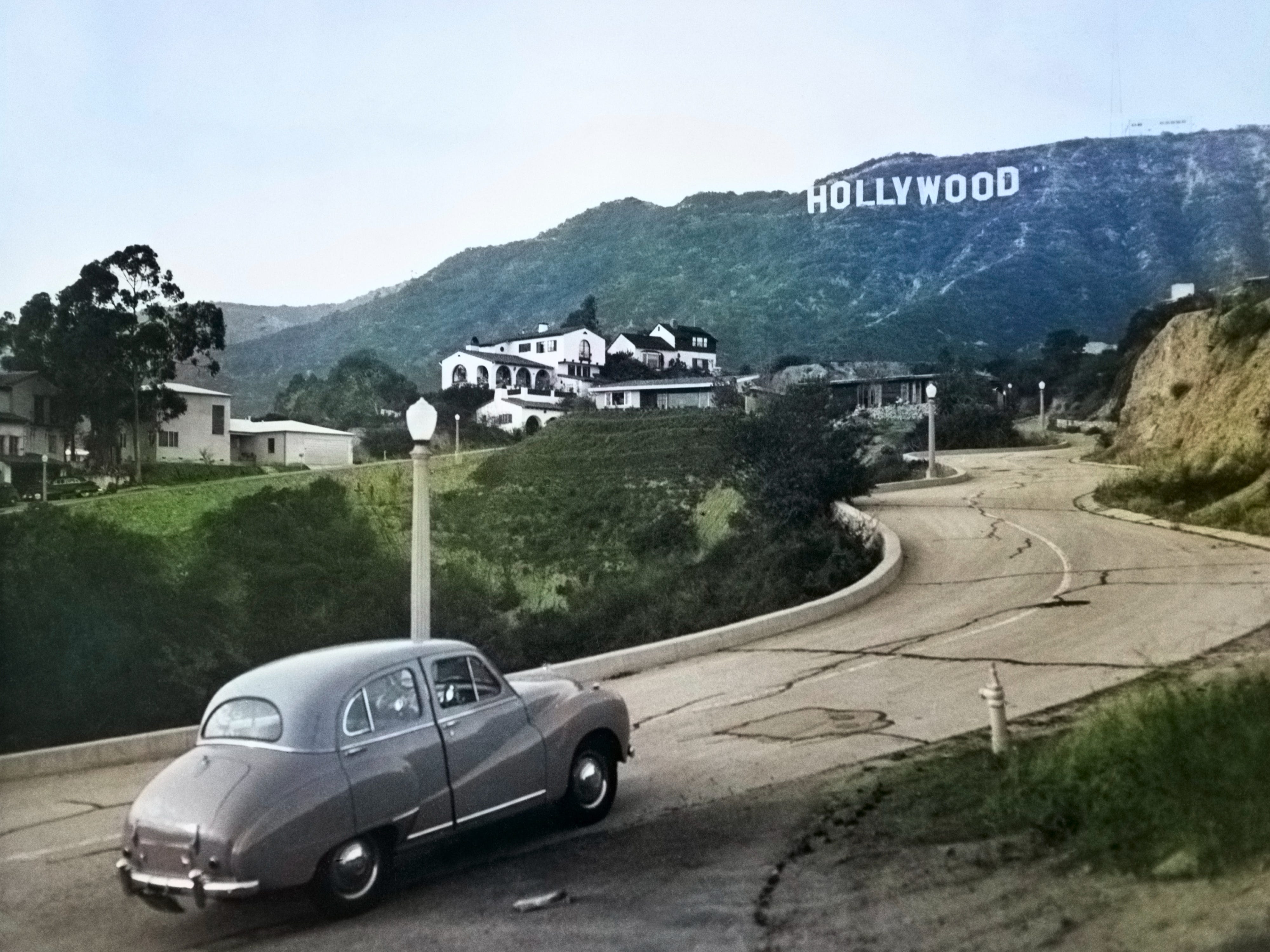 Ein Austin-Auto aus den 1950er Jahren fährt eine Straße in den Hollywood Hills hinauf, mit dem Hollywood-Schild in der Ferne in Los Angeles, Kalifornien.