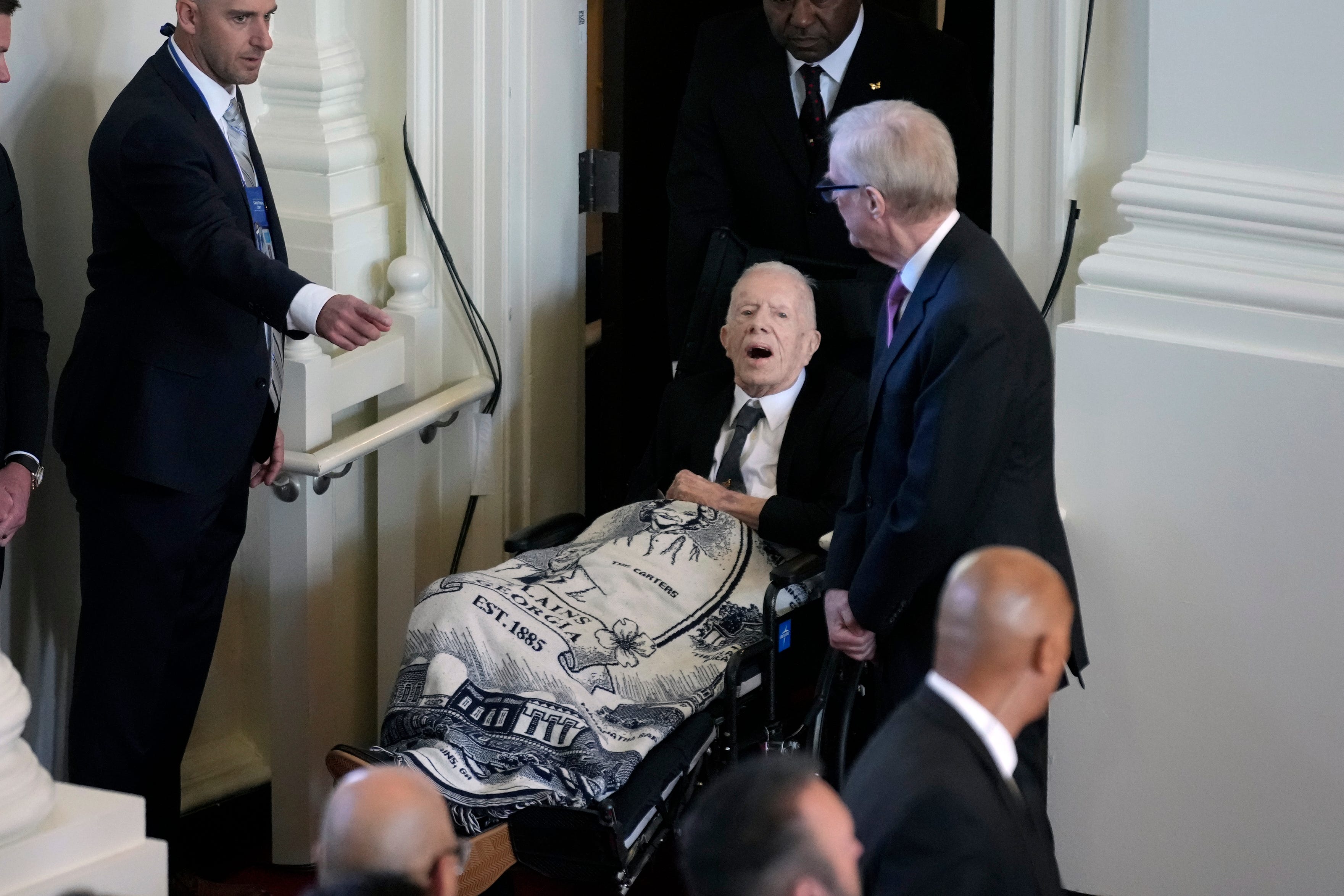 Ein Foto von Jimmy Carter, wie er im Rollstuhl zum Gottesdienst für seine Frau in eine Kirche begleitet wird.