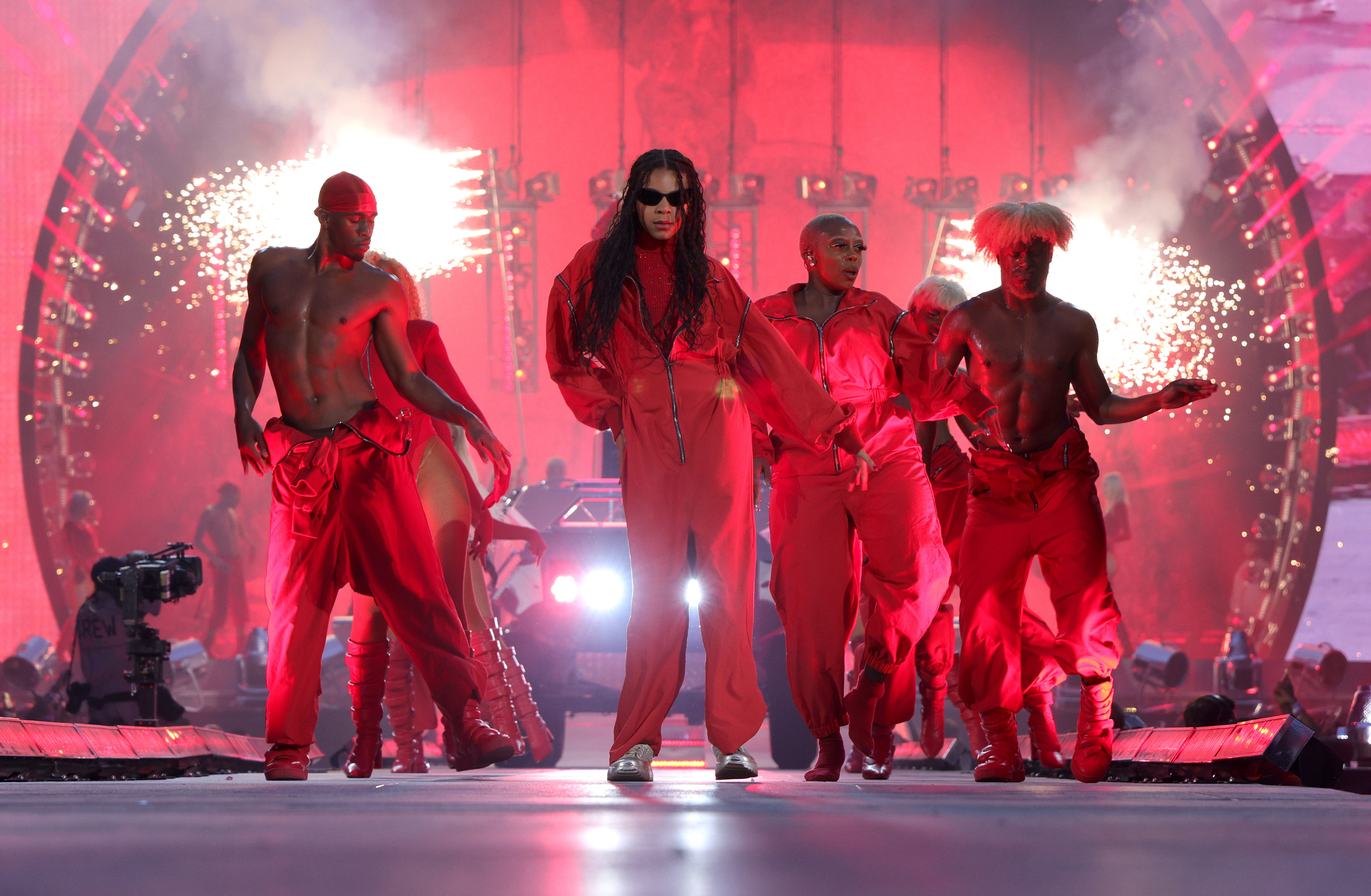 Blue Ivy in einem roten Overall und Turnschuhen, umgeben von fünf Tänzern mit ähnlichen Outfits, springt, während sie auf einer großen roten Bühne steht