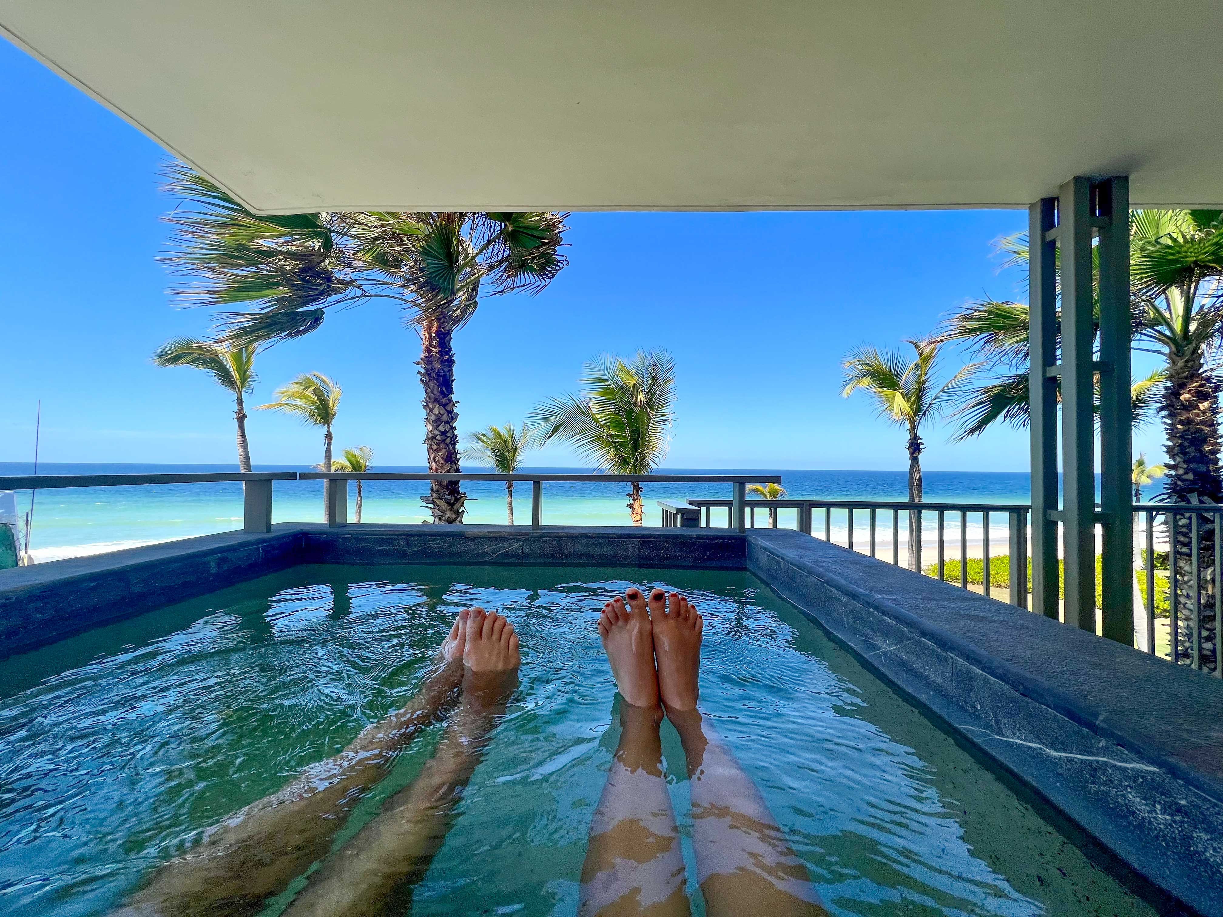 Zwei Paar Beine und Füße ragen aus einem Tauchbecken auf einem Balkon mit Blick auf einen tropischen Strand.